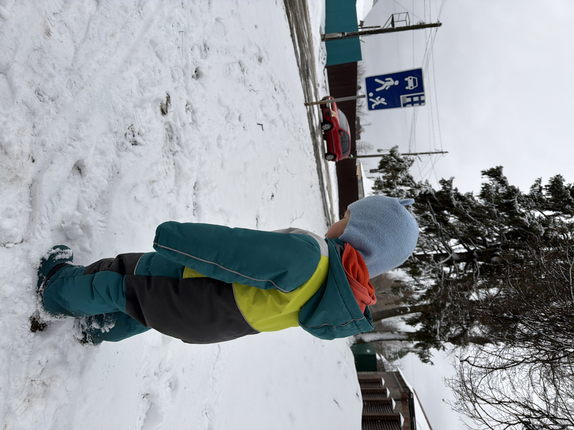 A young child dressed in a blue hat, a green and yellow jacket, and green boots stands in the snow. In the background is a street with a red car parked on the side and a blue sign with a pedestrian and a car depicted on it. The presence of the blue sign, a red car, and the presence of snow suggests this is Chaikovsky, Russia. A snow-covered tree is also visible to the right.