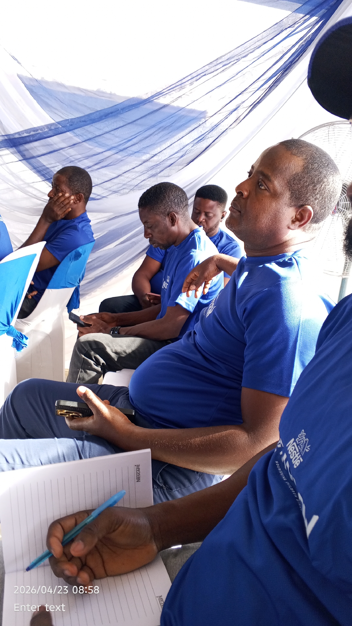 This media file captures a group of men attending an event or meeting, likely in Tarauni, Nigeria. The setting features white and blue draped fabric overhead, suggesting an indoor or tented venue.
The primary subjects are several adult men, all dressed in matching royal blue short-sleeved t-shirts. In the foreground, a man is looking upwards and to the right, appearing attentive or contemplative. His blue t-shirt prominently displays the white Nestlé logo and the partially visible slogan "NOURISHING CHILDREN". Adjacent to him, another man is focused on a mobile phone held in both hands. Further back, other participants are seated, with one resting his hand on his chin, seemingly listening. A white electric fan is partially visible in the upper right, suggesting the need for cooling.
In the bottom left foreground, hands are visible holding a mobile phone and a blue pen, actively writing in an open, lined notebook. The notebook pages are branded with "NESCAFÉ". The activity suggests a session where participants are observing, taking notes, or engaging with their devices.
Visible text includes the date and time "2026/04/23 08:58" in the bottom left corner, "Enter text" (likely a camera placeholder), "NESCAFÉ" on the notebook, and the "Nestlé" logo with "NOURISHING CHILDREN" on a t-shirt. The coordinated attire and branded items point towards a corporate or organized gathering.