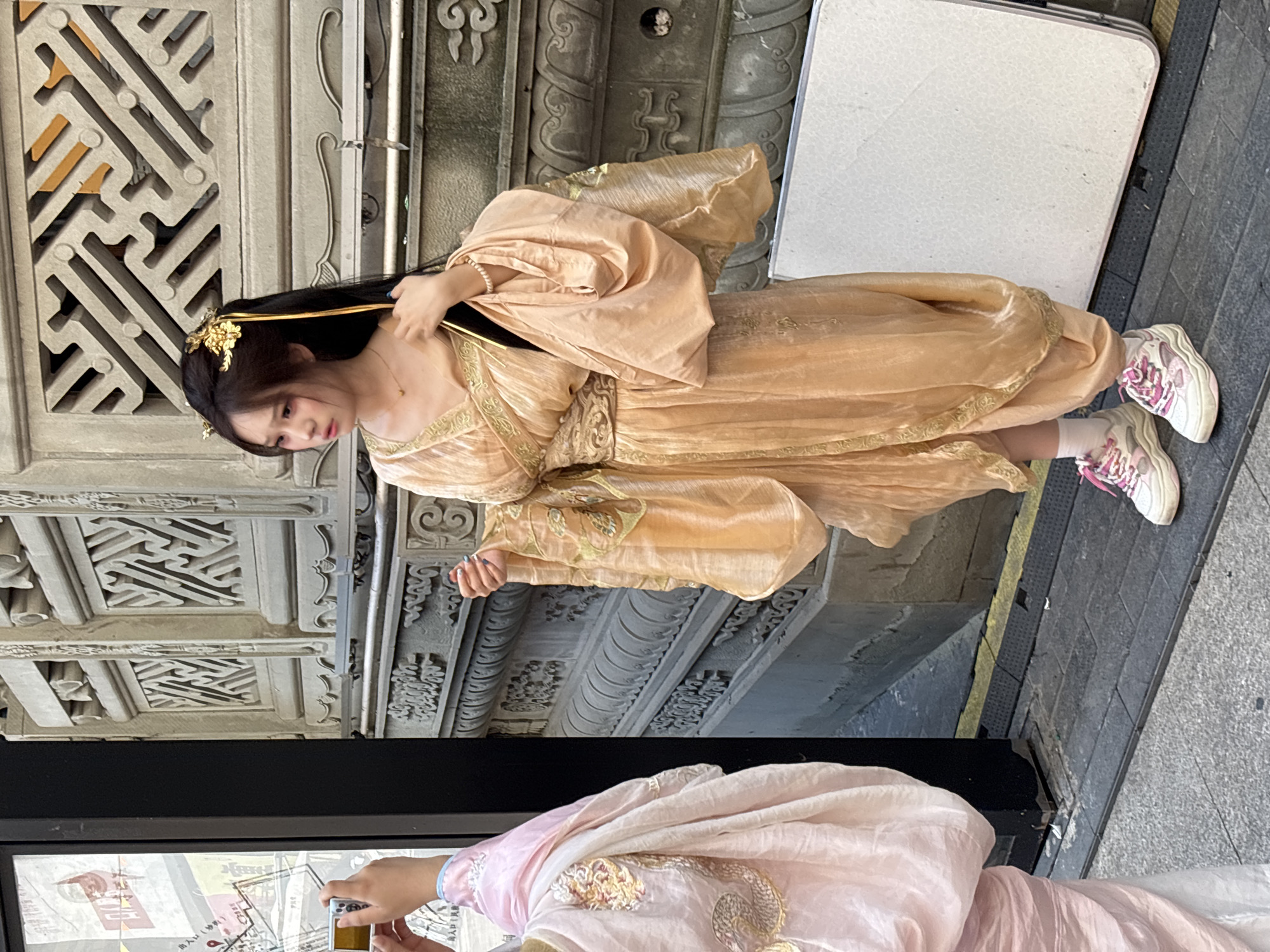 In Yuzhong District, China, a young East Asian woman stands in front of an intricately carved stone wall. She is wearing a traditional Chinese-style peach-colored satin dress with wide, flowing sleeves and gold trim. Her hair is long and dark, adorned with gold hairpins and a gold ribbon that drapes down her back. She also wears a delicate gold necklace and a pearl bracelet. Her feet are shod in white sneakers with pink laces. To her left, another person, partially visible, is dressed in a pale pink robe with gold embroidery and is holding a smartphone, seemingly taking a photograph of the woman in the peach dress. The background is a weathered, gray stone wall with ornate carvings and latticework. A white table is visible in the lower right corner of the frame, and the ground is paved with gray stones and dark, textured tiles. A black pillar is to the left of the frame, next to a display with Chinese text.