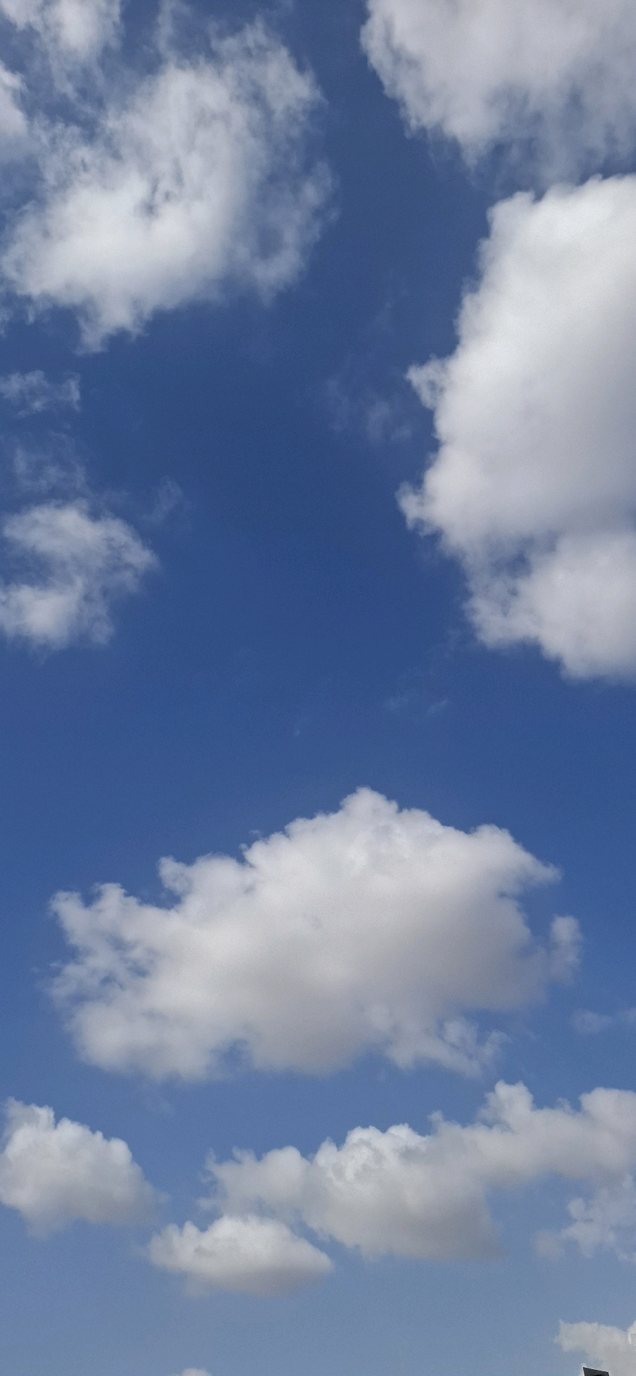 The image shows a clear blue sky with scattered white cumulus clouds. The clouds are varied in size and shape, with some appearing puffy and dense, while others are wispy and spread out. The lighting suggests it is daytime, with the sun likely high in the sky, illuminating the tops of the clouds. There are no visible man-made structures or natural landforms in the frame, indicating an unobstructed view of the sky. The perspective is from ground level, looking upwards. The image is likely taken in Kiryat Gat, Israel, given the context provided.