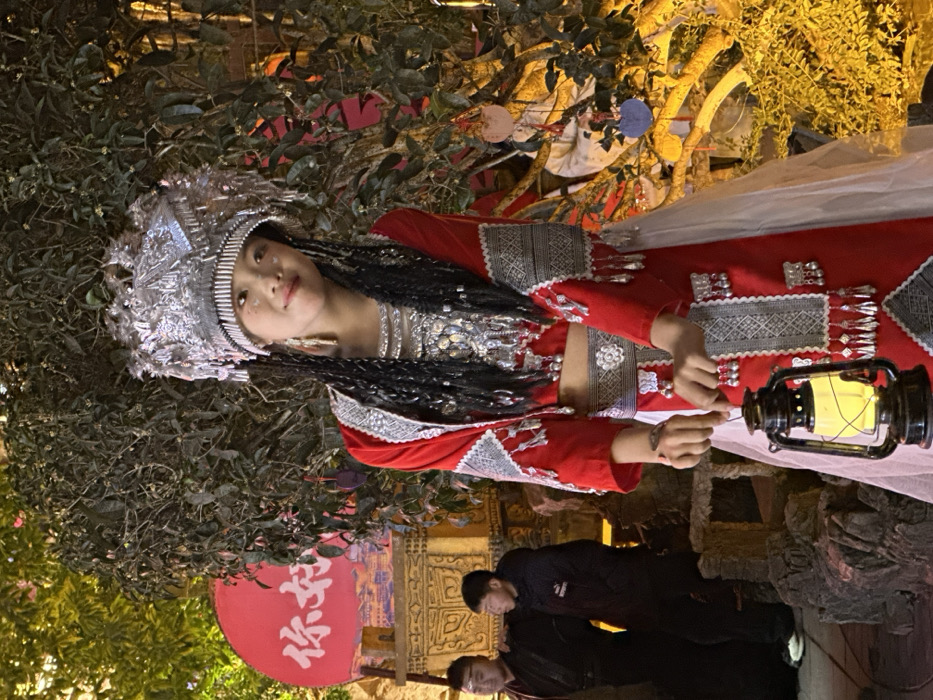 An outdoor night scene in Yuzhong District, China, depicts a young female standing centrally, attired in elaborate traditional ethnic costume. She wears a large, ornate silver headdress with intricate patterns and fringe, a red cropped long-sleeved top with silver embroidery and dangling ornaments, a matching red skirt with similar embellishments, and a silver belt. Her dark hair is styled in multiple braids adorned with silver accents. She holds a lit black lantern with a yellow glow in both hands at waist height and looks directly forward with a slight smile.
To her left, partially obscured in the mid-background, are two males. The leftmost male wears a dark, possibly traditional robe. The male to his right wears a dark puffer jacket with a white Puma logo on the left chest.
The background consists of dense green foliage illuminated by warm yellow artificial light. A large, circular red sign with white Chinese characters "你和" (Nǐ hé) and a stylized cityscape silhouette is partially visible behind the males. Ornate, carved stone or wooden structures are visible in the left mid-ground, and small, colorful decorative elements hang from the foliage on the right. The overall lighting suggests a nighttime setting within a culturally themed area.