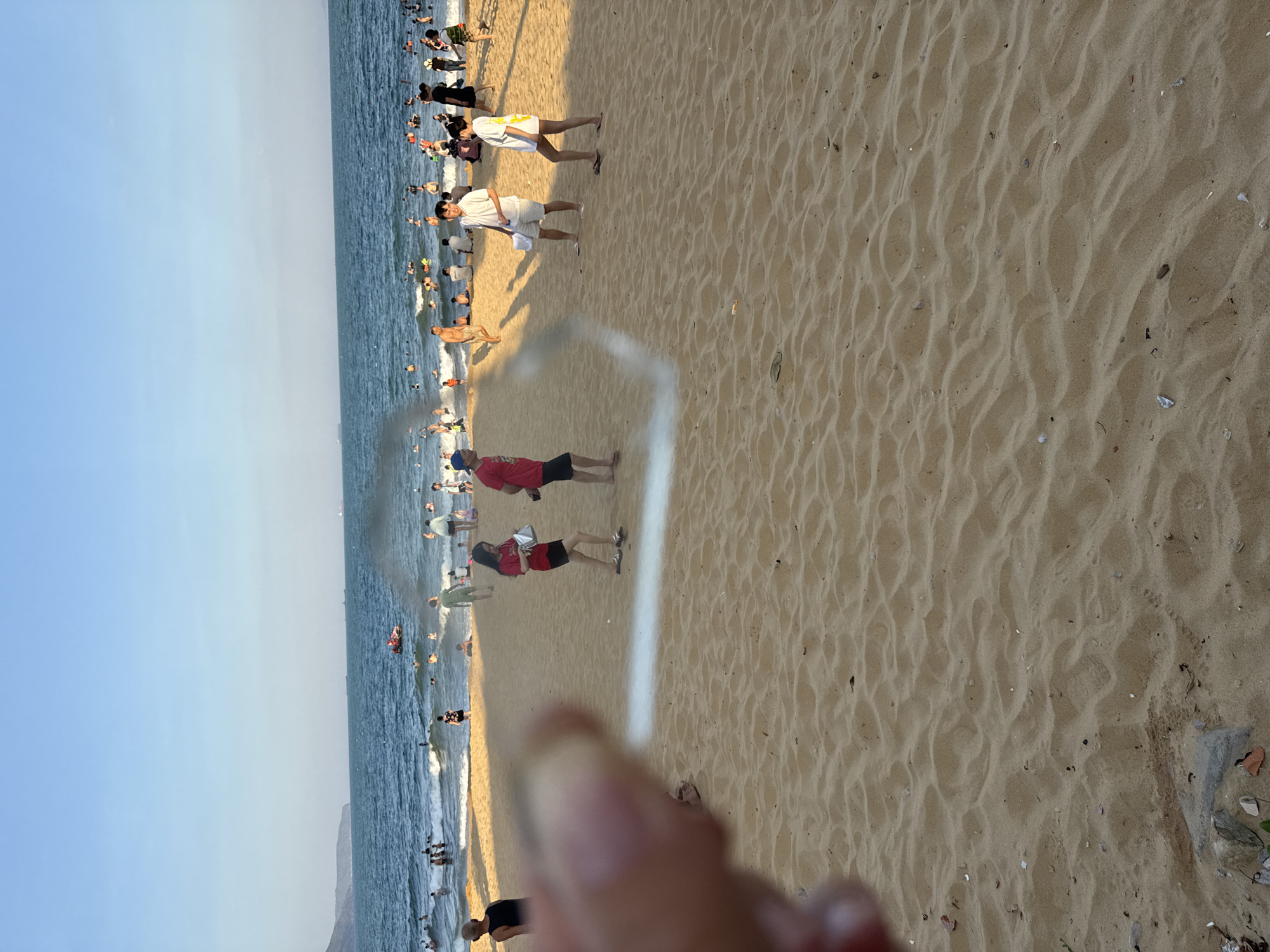 The image captures a vibrant, sun-drenched beach scene at South Quy Nhon Ward, Vietnam, likely during the late afternoon given the warm, golden light and long shadows cast on the sand. A diverse crowd of people are enjoying a day at the beach. Many are in the shallow ocean water, swimming and playing in the gentle waves. Others are on the sandy shore, walking, socializing, or relaxing.
The beach is characterized by its golden sand, which shows ripples from the receding tide and scattered debris like small shells and pebbles. The ocean is a deep blue, with white foam cresting on the incoming waves. The sky above is a clear, pale blue, indicating fair weather.
Several individuals are prominently featured. Near the water's edge, a man in a red shirt and black shorts stands facing away from the camera. Further up the beach, two people in red shirts and dark shorts are walking. A person in a white shirt and shorts is also visible walking on the sand. Further back, a cluster of people are wading in the water, and many more are scattered throughout the scene, indicating a popular recreational spot.
There are no visible signs or text within the image. The overall mood is one of leisurely enjoyment and a typical summer day at a busy beach. The perspective is from someone standing on the sand, looking towards the ocean and the crowded shoreline. A blurry foreground suggests the presence of the photographer's hand or an object in close proximity to the lens.