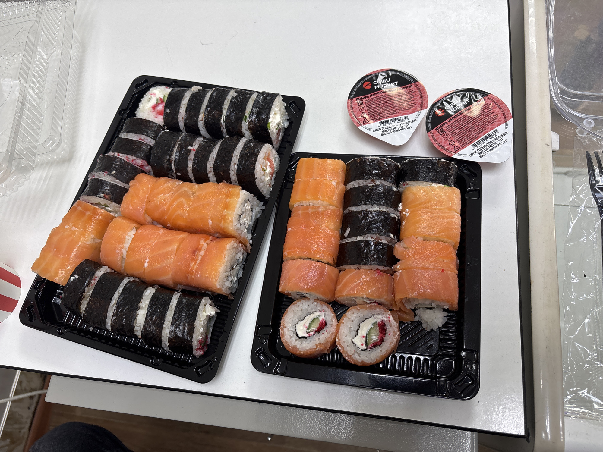 In Фролы, Russia, on a white surface, two black plastic trays of sushi are presented. The upper tray contains several rolls of sushi, some wrapped in nori seaweed and filled with rice, cream cheese, and visible red roe, while others are topped with slices of bright orange salmon. The lower tray features a similar arrangement of sushi rolls, with a focus on those topped with salmon and some with visible cream cheese and red roe. Next to the lower tray are two small, round, red-lidded containers of soy sauce or a similar condiment. The background includes a hint of a person's dark clothing and a partially visible clear plastic food container. The overall scene depicts prepared food ready for consumption.