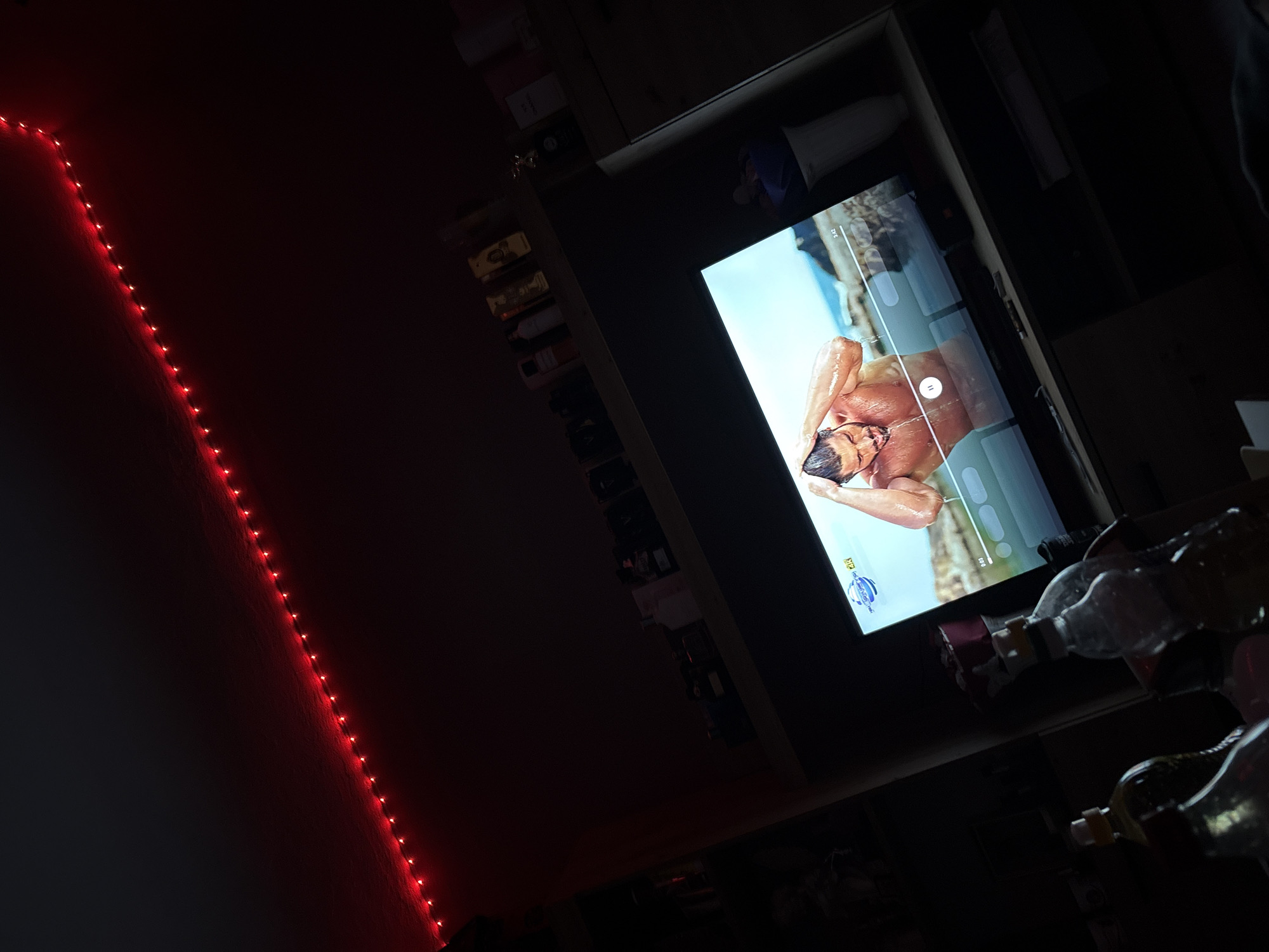 The image depicts a dimly lit room with a television displaying a man in an aquatic setting, likely a movie or video. A string of red lights is visible along the wall to the left. A shelf to the right of the television holds a variety of items, including bottles and containers. The photo was not taken in Kazichene, Bulgaria.