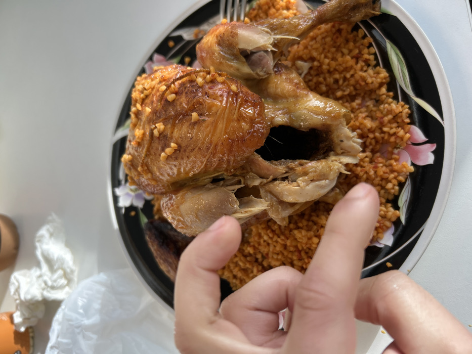 The image depicts a partially consumed roasted chicken served on a bed of orange-colored bulgur or similar grain, situated on a black circular plate with a floral design. The chicken, golden-brown and exhibiting signs of internal dissection, is topped with small, light-colored颗粒, possibly nuts or spices. A left human hand, with fingers slightly curled, reaches towards the chicken, positioned centrally within the frame. A silver fork is partially visible on the right side of the plate. The plate rests on a white, unadorned surface, likely a table. In the blurred background, several domestic items are visible, including a crumpled white plastic bag, a piece of white tissue, and parts of an orange cup and a brown object. The scene occurs indoors, within the city of Illkirch-Graffenstaden, France.