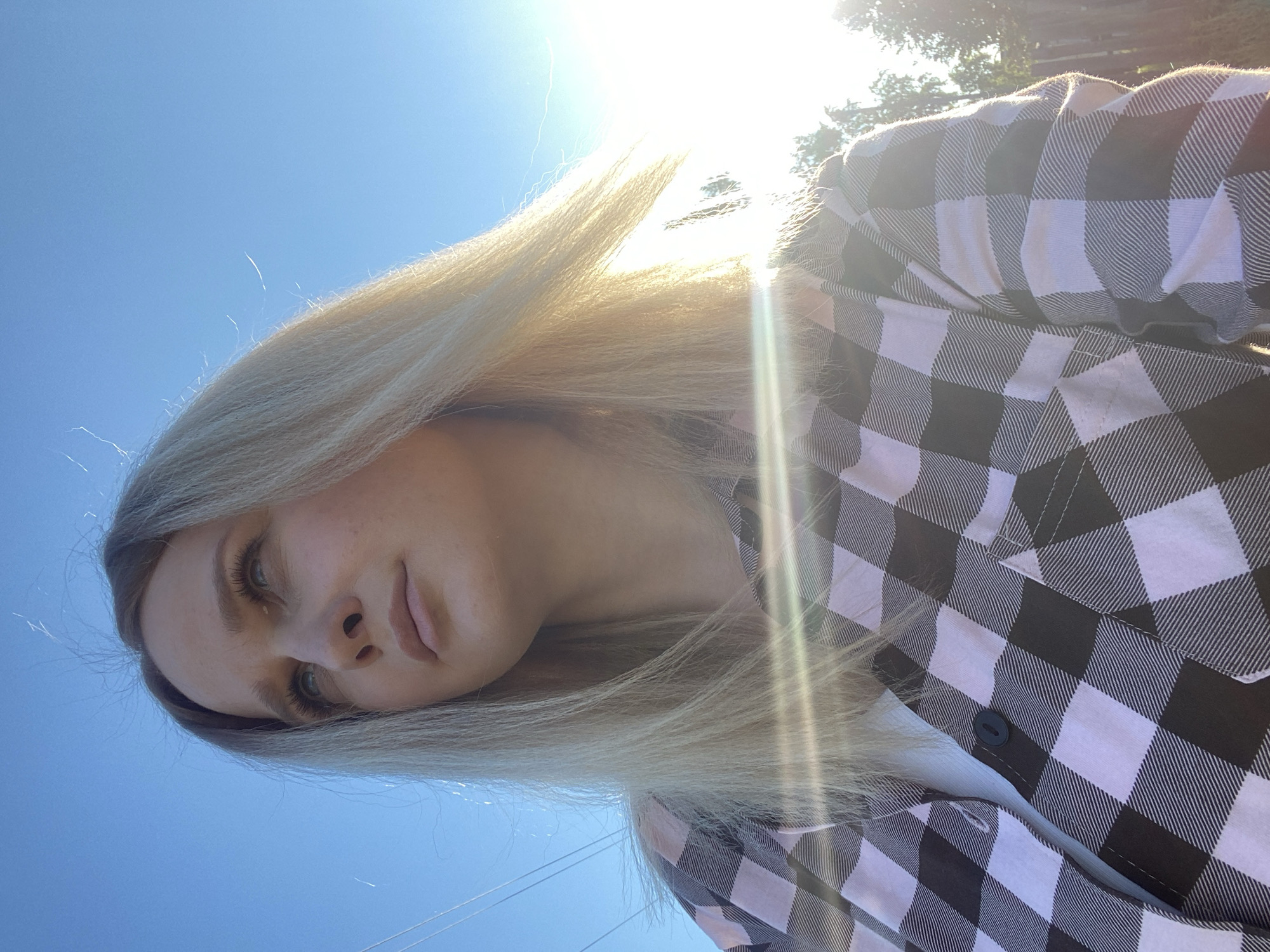 An upward-angled outdoor portrait depicts a person with long, straight, blonde hair, taken under a clear, bright blue sky. The image is rotated 90 degrees counter-clockwise. Strong sunlight from behind and above the subject creates significant lens flare and backlighting, causing the hair to glow and individual strands to stand out. The person's face is partially shadowed by the backlighting but details of their light-colored eyes, nose, and lips are visible, revealing a neutral to slightly pensive expression.
The person is wearing a black and white plaid shirt. The pattern features prominent black and white squares, with the white squares having fine diagonal lines for texture. A single black button is visible on the shirt. In the background, faint outlines of green foliage from trees can be seen through the strong sunlight. The overall setting suggests a sunny daytime environment in Kauchuk Stantsiya, Russia. There is no visible text in the image.