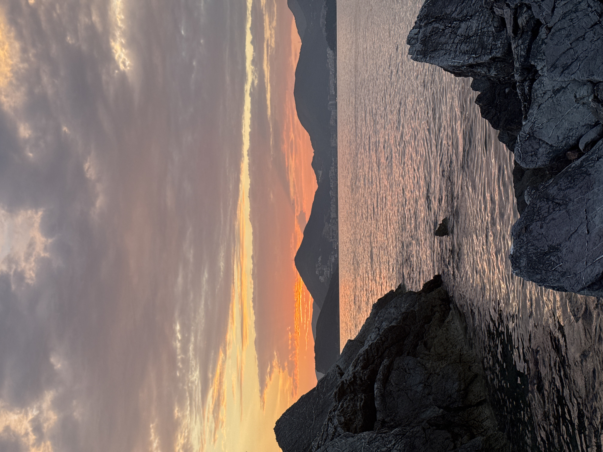 The image depicts a sunset over the Adriatic Sea, viewed from a rocky coastline near Sveti Stefan, Montenegro. In the foreground, dark, textured rocks dominate the left and right sides of the frame, with a small inlet of calm water on the left. The sea, reflecting the sunset's colors, occupies the middle ground, extending towards a distant mountain range. These mountains are silhouetted against the vibrant sky, with a scattering of lights indicating a settlement on their slopes. The sky itself is filled with a gradient of colors, from warm oranges and yellows near the horizon to cooler purples and grays at the top, with scattered, wispy clouds. No people or animals are visible in the scene. The overall impression is one of natural beauty and tranquility at dusk.