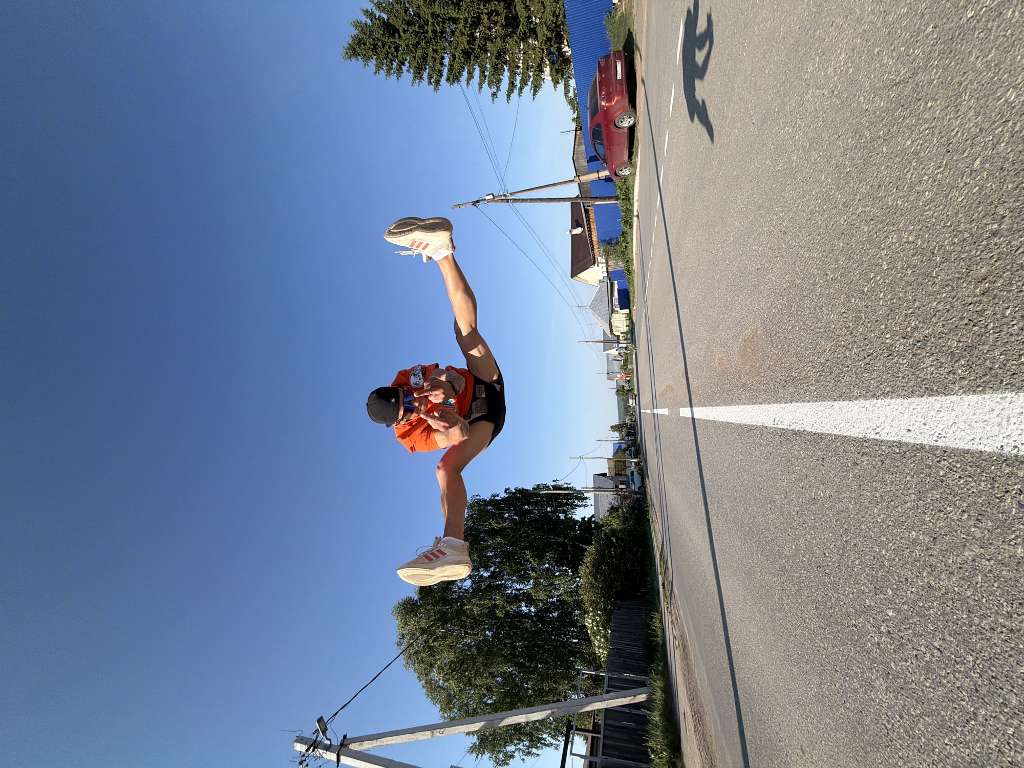 A person is in a full split in the air, wearing an orange t-shirt, black shorts, a black baseball cap, and white athletic shoes with pink stripes. They appear to be holding a cell phone. The location is a paved road with a white dividing line, flanked by vegetation and buildings. There is a red car parked on the roadside. Power lines and poles are also visible. The sky is clear and blue. The location is the city of Chaikovsky, Russia.