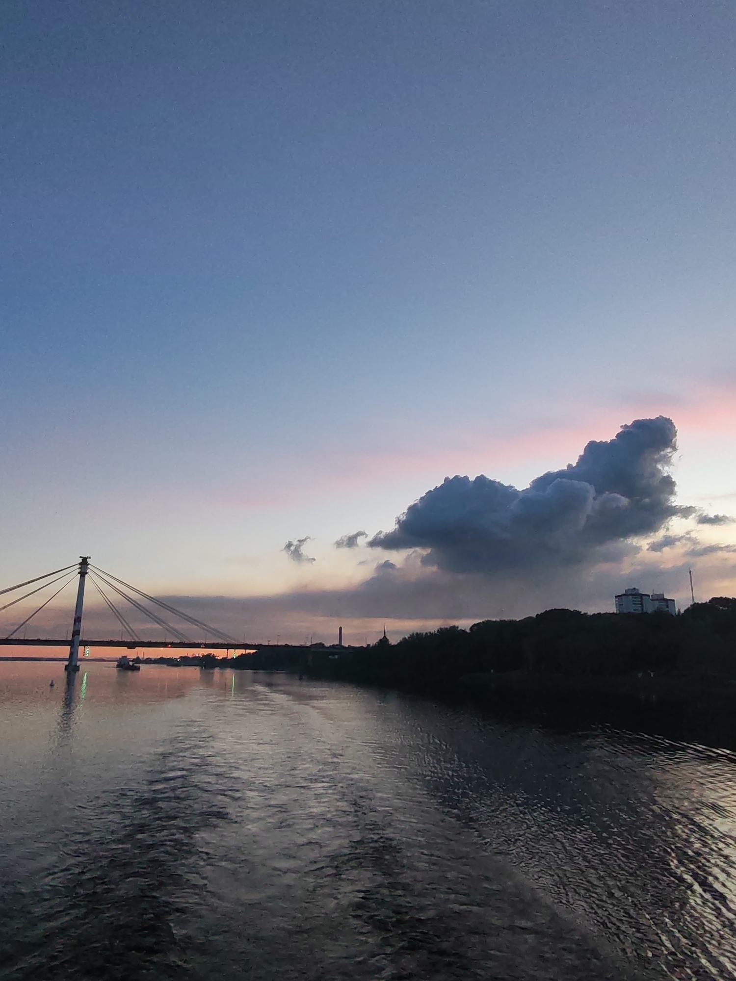 The image depicts a river scene at dusk in Cherepovets, Russia. A cable-stayed bridge spans the river on the left, with red and white painted support structures. The water surface reflects the muted light. On the right, the riverbank is lined with trees, obscuring most buildings, but a multi-story structure is visible in the distance. A prominent, dark cloud formation hangs above the tree line, with pink hues in the sky above it. The sky transitions from blue at the top to orange/pink near the horizon. There is boat in the river.