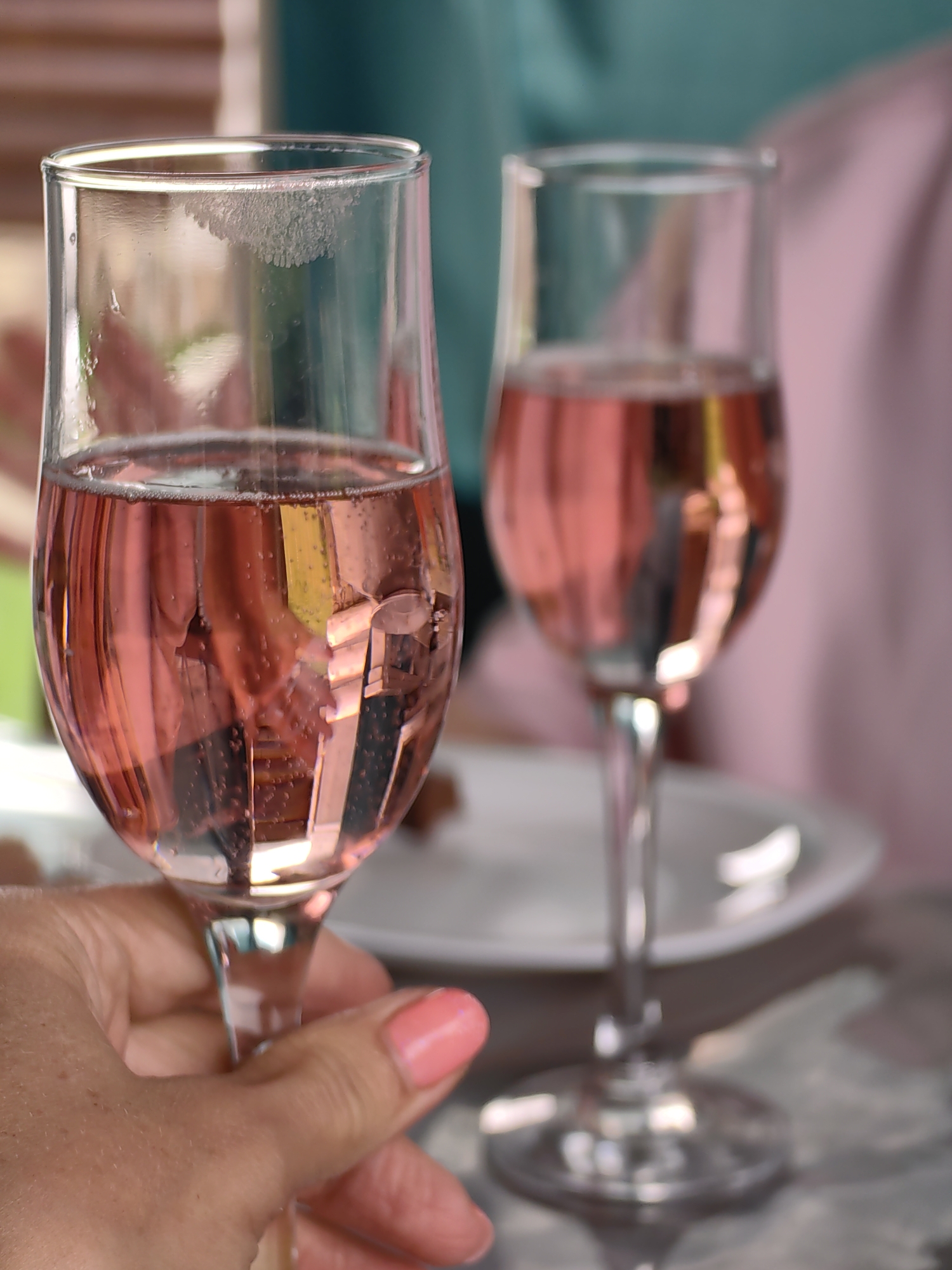 A close-up, eye-level shot captures two flutes filled with pink champagne. The left flute is held in the foreground by a hand with bright pink nail polish. A faint lipstick mark is visible on the rim of this flute. In the background, the second flute stands on a table, with two blurred figures, one in a teal top and another in a pink garment, also blurred, are visible behind the flutes. The setting appears to be a celebratory gathering, possibly in a restaurant or at home. The location in the city of Myadzyel, Belarus cannot be determined from the image.