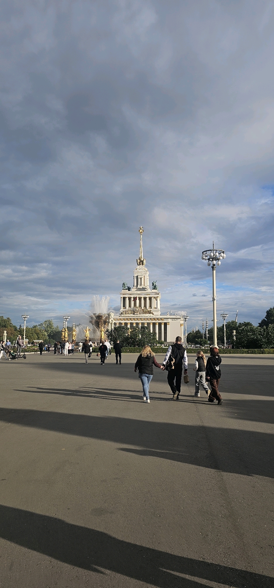 The image depicts an outdoor scene in Moscow, Russia, likely at VDNKh (Exhibition of Achievements of National Economy). In the background, a grand, ornate building with a golden spire and classical architectural elements stands prominently. In front of this building, a large fountain sprays water into the air, with golden statues visible around its base. Several people are present in the scene, walking across a paved plaza. In the foreground, a family or group of individuals, including adults and children, are walking away from the camera, some holding hands. Other individuals and a person on an electric scooter are visible further back in the plaza. The sky is partly cloudy with dramatic cloud formations. The overall atmosphere suggests a public gathering or a tourist attraction in a city setting.