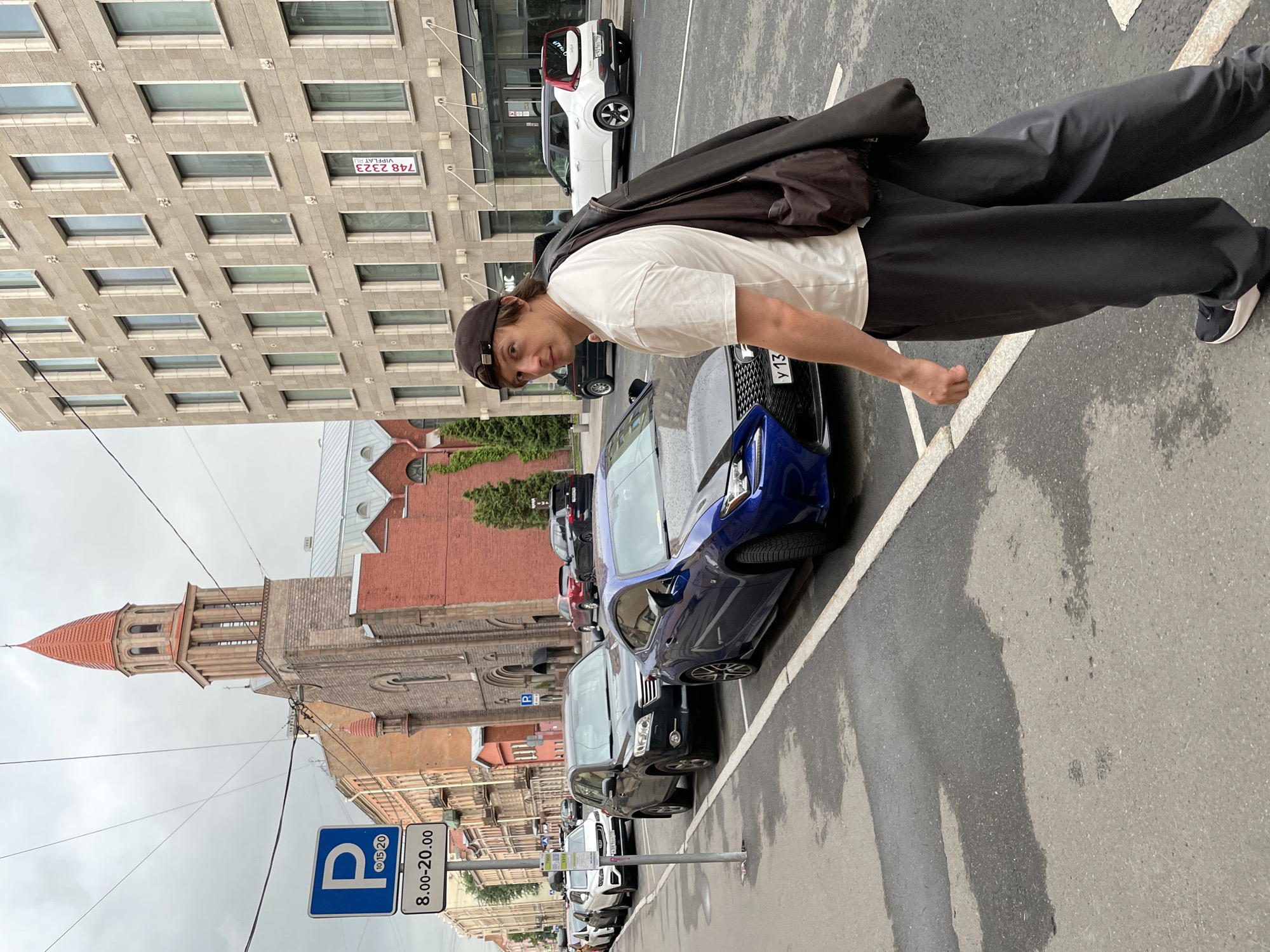 A man wearing a white t-shirt, black pants, a dark baseball cap, and sneakers walks away from the camera on a wet street in Saint Petersburg, Russia. He carries a dark jacket slung over his right shoulder and a dark bag. He is positioned in the foreground, to the right of a blue Lexus RC F. Behind the Lexus, other cars are parked along the street, including a black SUV, a white Kia Soul, and other vehicles. A red brick church with a tall, slender tower is visible in the background, to the left. A modern, multi-story building with many windows stands to the right of the church. A blue parking sign with a 'P' and time restrictions is posted on the left side of the street. The weather appears overcast.