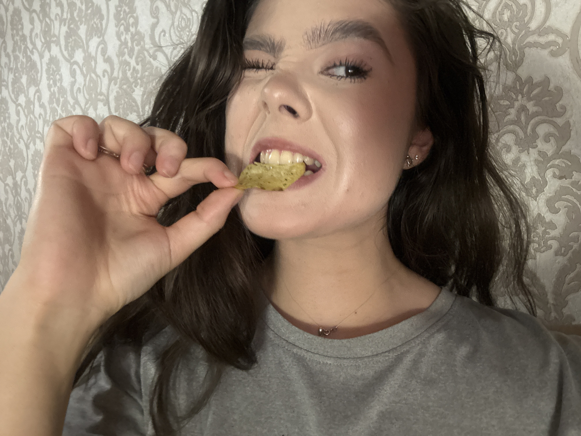 The image is a close-up, chest-up shot of a young woman with dark, wavy hair and fair skin. She is wearing a gray t-shirt and a delicate necklace with two small rings. Her left eye is closed in a wink, while her right eye is open, looking towards the camera. She is holding a single, thin, rectangular-shaped chip, possibly a cracker or a vegetable chip, between the thumb and index finger of her left hand, bringing it to her mouth. Her teeth are slightly parted, and her lips are glossed. The background is a textured wallpaper with a light, ornate floral pattern. The lighting appears to be artificial and somewhat dim, casting shadows that highlight the contours of her face and hand. The overall impression is casual and playful. There is no discernible indication of the city of Zhovte, Ukraine within the image itself.