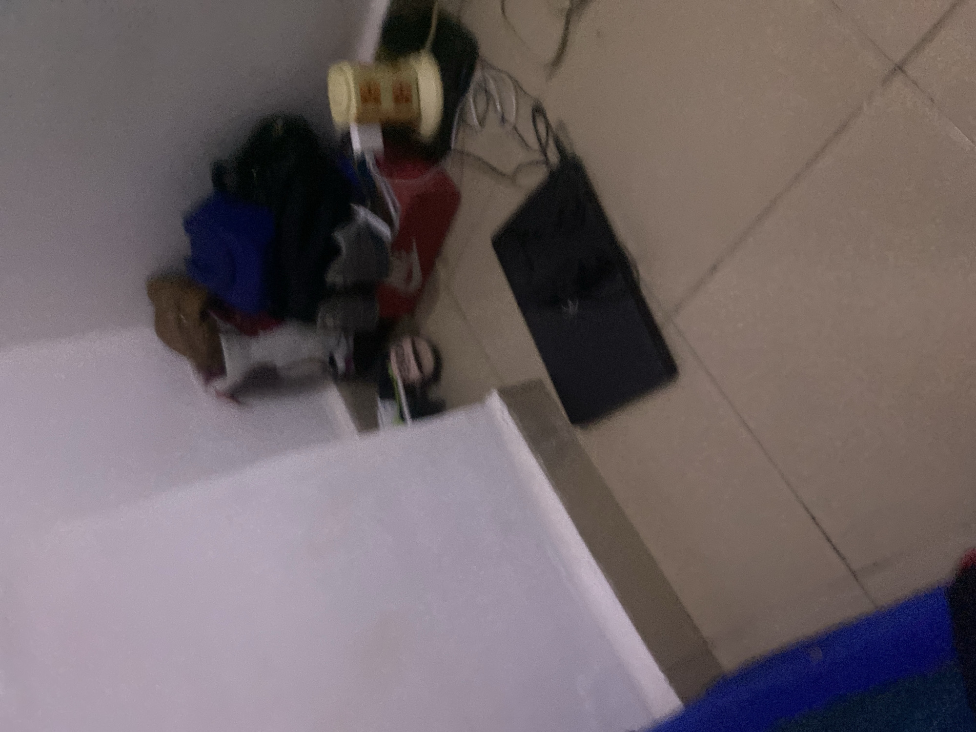 The image depicts a cluttered corner of a room with tiled flooring. A white wall forms the background, with a beige skirting board. In the corner, several items are piled up, including a blue garment, a black bag, and what appears to be a red Nike shoe box. A yellow cylindrical object with orange markings is also visible. On the floor in the foreground, a black laptop is placed open, with visible wires connected to it. The overall impression is one of disarray, with objects scattered rather than neatly stored. There is no indication of the specific location within Howayi, Nigeria, as the image only shows an interior scene.