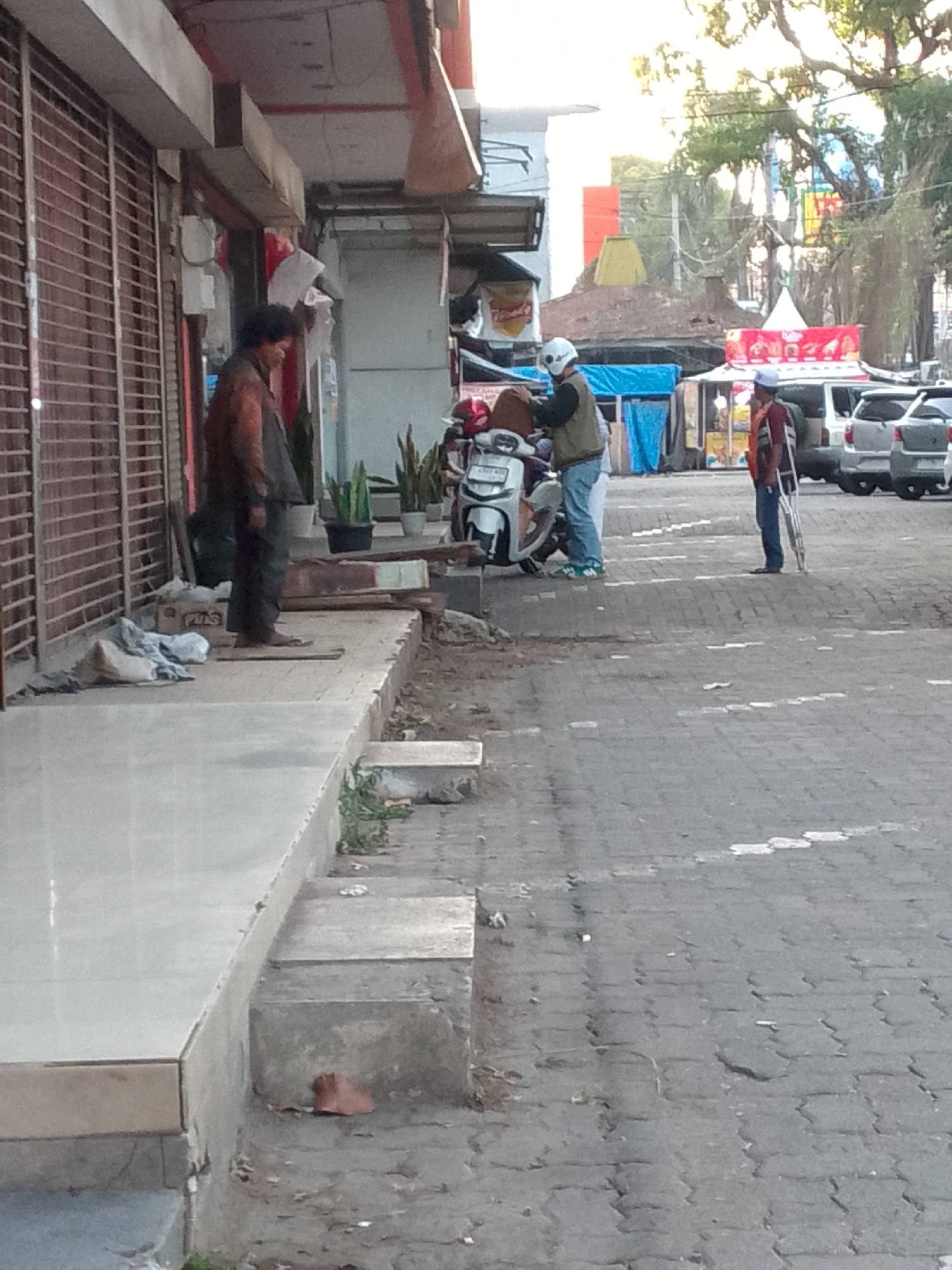 In Kertamulya, Indonesia, a street scene unfolds with several individuals and a parked motorcycle. To the left, a person with dark, curly hair wearing a brown jacket and dark pants stands on a raised sidewalk near a storefront with closed metal shutters. Adjacent to this storefront, a person in a light-colored shirt and blue pants, wearing a white helmet, is seated on a motorcycle. Nearby, another individual, also wearing a white helmet, is standing. Further to the right, a person in a maroon shirt and blue pants, using crutches, stands on the paved street. Behind them, several vehicles, including cars and SUVs, are parked. The environment appears to be an urban street with a mixture of storefronts and paved surfaces, under natural daylight with some trees visible in the background.