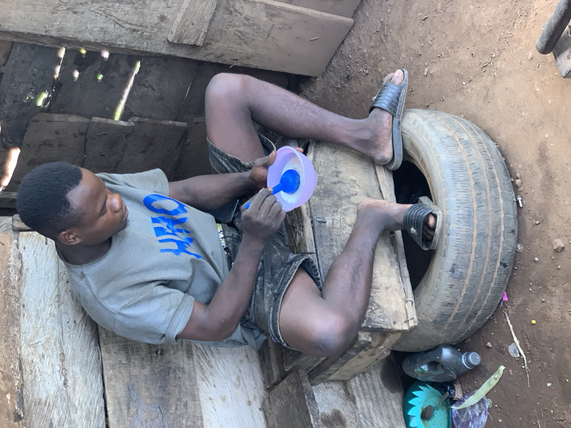 In Gbeleko, Nigeria, a young Black man is seated on a makeshift wooden structure composed of stacked planks, which is itself perched atop a worn vehicle tire. He is wearing a grey t-shirt with blue lettering and dark denim shorts. He is holding a light purple bowl with a blue spoon, appearing to be eating or preparing food. His feet are bare except for black slide sandals. The surrounding environment consists of rough, wooden walls, suggesting a rustic or unfinished structure. Debris, including plastic bottles and what appears to be a green plastic item, is visible on the dirt ground around the tire. The scene is illuminated by natural light filtering into the structure.