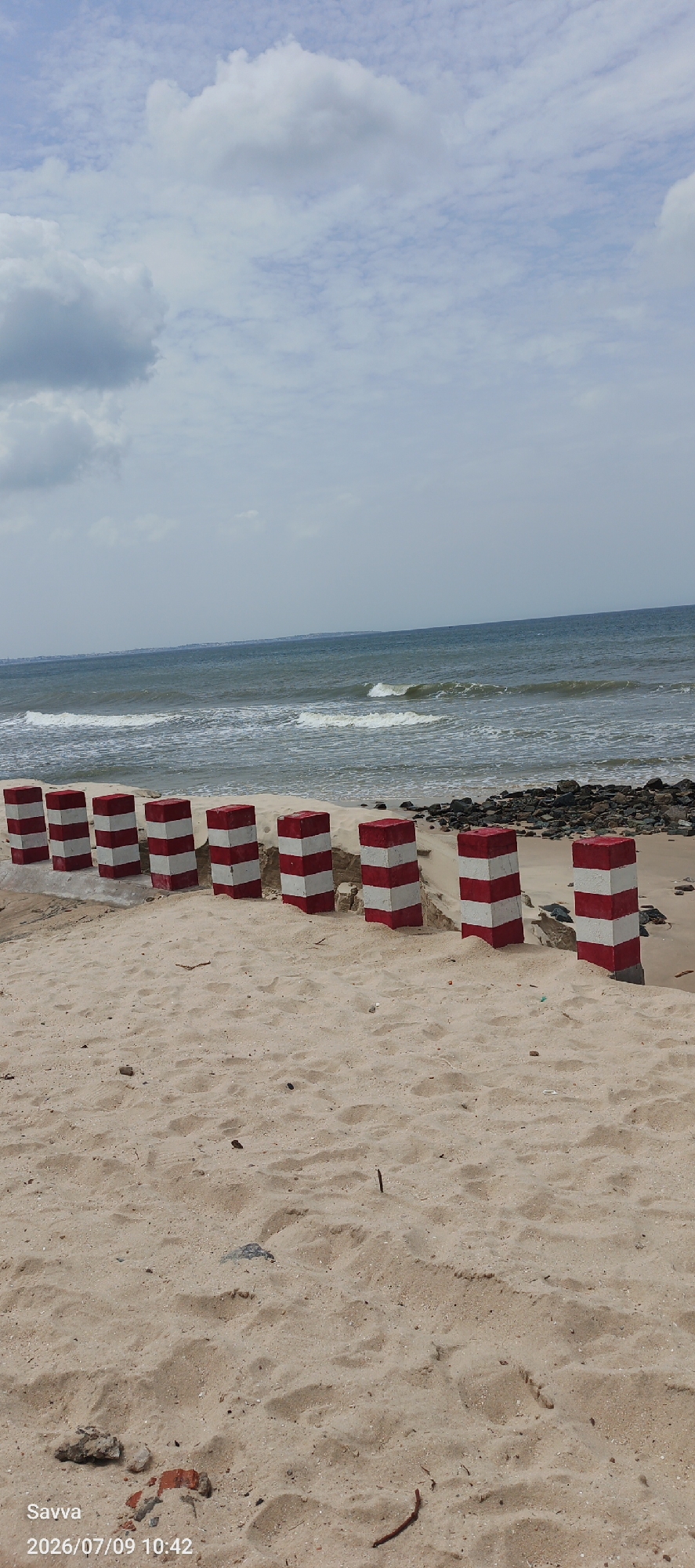 The image depicts a serene beach scene in Mũi Né, Vietnam. In the foreground, a sandy beach stretches out, with a line of red and white striped concrete bollards marking the edge of the shore. These bollards appear to be part of a barrier or pathway.
Beyond the sand, the ocean is visible, with gentle waves rolling towards the shore. The water is a deep blue, and the sky above is partially cloudy, with scattered white and gray clouds. The overall atmosphere is peaceful and natural, characteristic of a coastal environment.