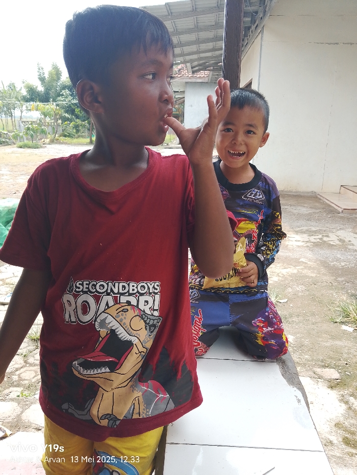 The image shows two young boys standing in front of a building in Anjatan, Indonesia (latitude: -6.419, longitude: 107.978). The boy on the left, wearing a red shirt with a cartoon dinosaur on it, is looking directly at the camera and is sticking his finger in his mouth. The boy on the right, wearing a multi-colored, patterned shirt, is looking at the boy on the left and smiling. He is holding a bag of chips in his right hand and is holding his left hand on his hip. The boys are standing in front of a cement building with a concrete floor. The floor is dirty and there are some cracks and holes in it. The background is a blurry image of trees and bushes. The image is likely taken on a sunny day.