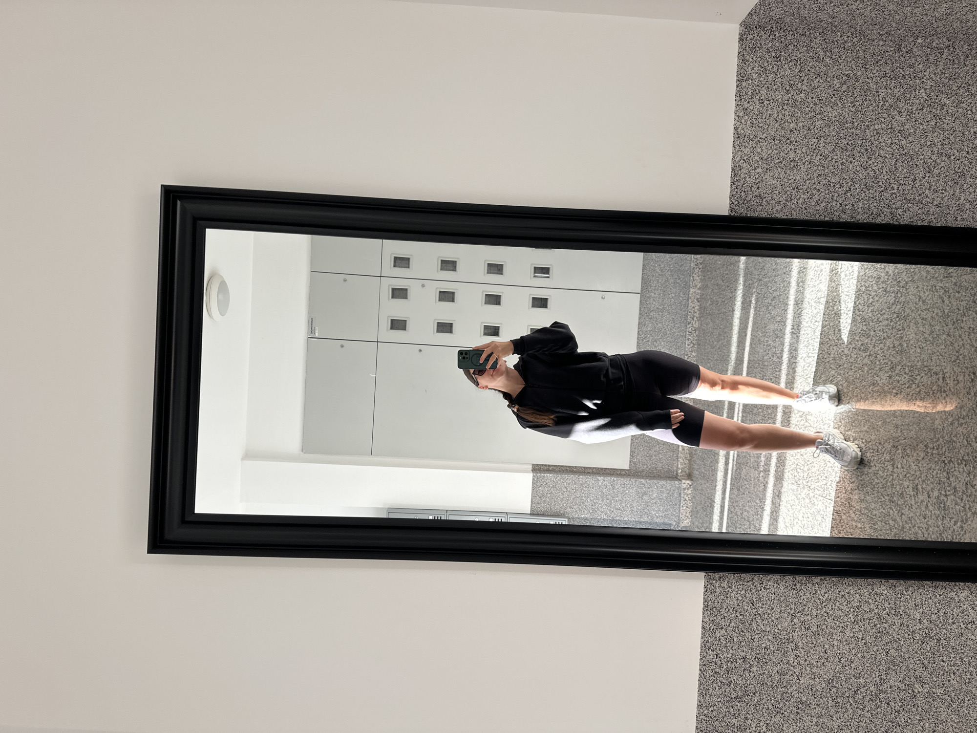 A person in black athletic wear is taking a mirror selfie in a hallway with a light gray wall on the left and a speckled gray wall on the right. The mirror has a thick black frame. The reflection shows lockers on the wall behind the person and sunlight coming through what appear to be windows or a door. The location of the image is a hallway of the school, International School of Montenegro in Budva, Montenegro.