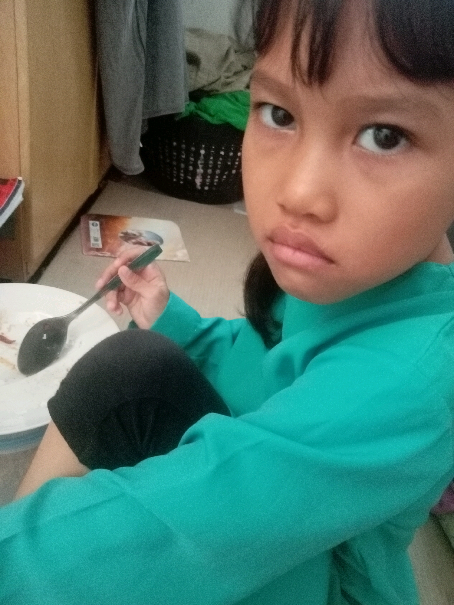 The image shows a close-up view of a young girl with dark hair, dark eyes, and light brown skin. She is wearing a teal long-sleeved shirt and black leggings. Her expression appears to be slightly unhappy or pensive, with her lips slightly pursed. She is holding a dark-colored spoon in her right hand, which is resting on a white plate. Part of the plate and some food residue are visible.
The background is cluttered. To the left, a wooden surface and a stacked item with red and white elements are visible. Further back, a black laundry basket with a white pattern is present, containing some green fabric. Behind the basket, grey pants are hanging, and a light-colored wall or furniture is visible. The floor appears to be tiled or carpeted in a neutral tone.
The setting suggests an indoor environment, possibly a home. The presence of laundry and personal belongings implies a domestic space. Without further context or external clues, it is not possible to definitively determine the specific location within Batam, Indonesia. The scene captures a candid moment, likely in a residential area of the city.