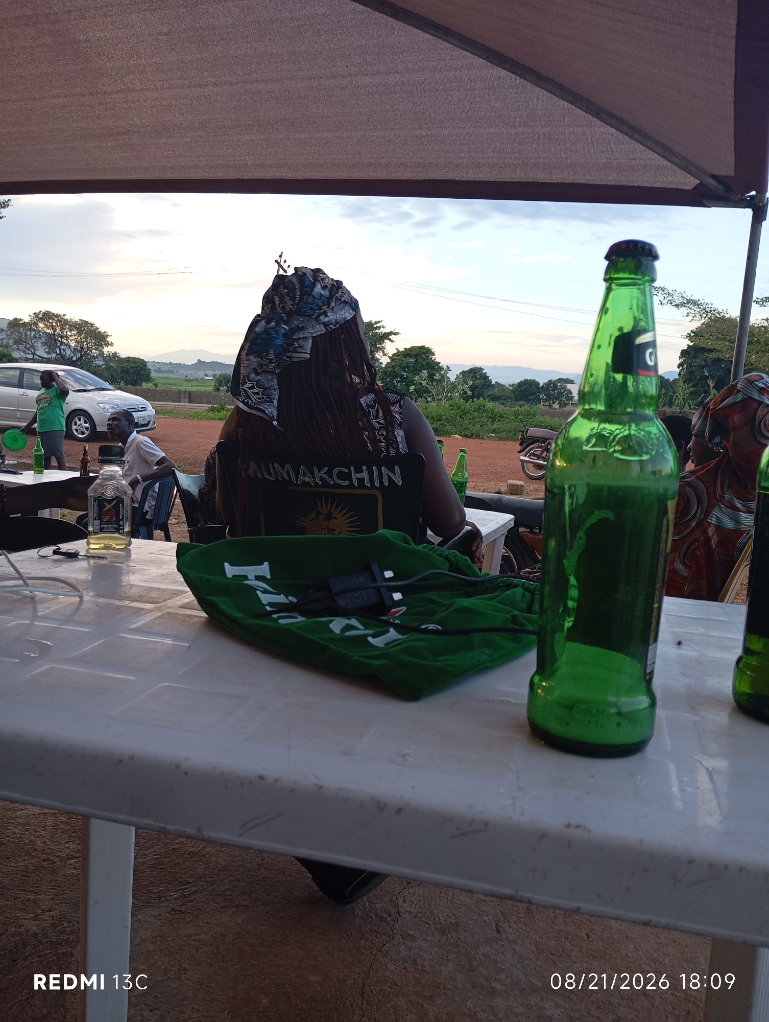 In Shendam, Nigeria, an outdoor scene unfolds beneath a beige canopy. A person, seen from behind, wears a dark shirt with "MAUMAKCHIN" printed on it and a patterned head covering. To their right, a green beer bottle stands prominently on a white plastic table. In the foreground, a green cloth is draped over the table, with a black electronic device resting on it. Other bottles and a clear liquor bottle are also visible on the table. In the background, a white car and a motorcycle are parked. The landscape features trees and distant hills under a cloudy sky, suggesting a rural or semi-rural setting. The timestamp "08/21/2026 18:09" indicates the time the photograph was taken.