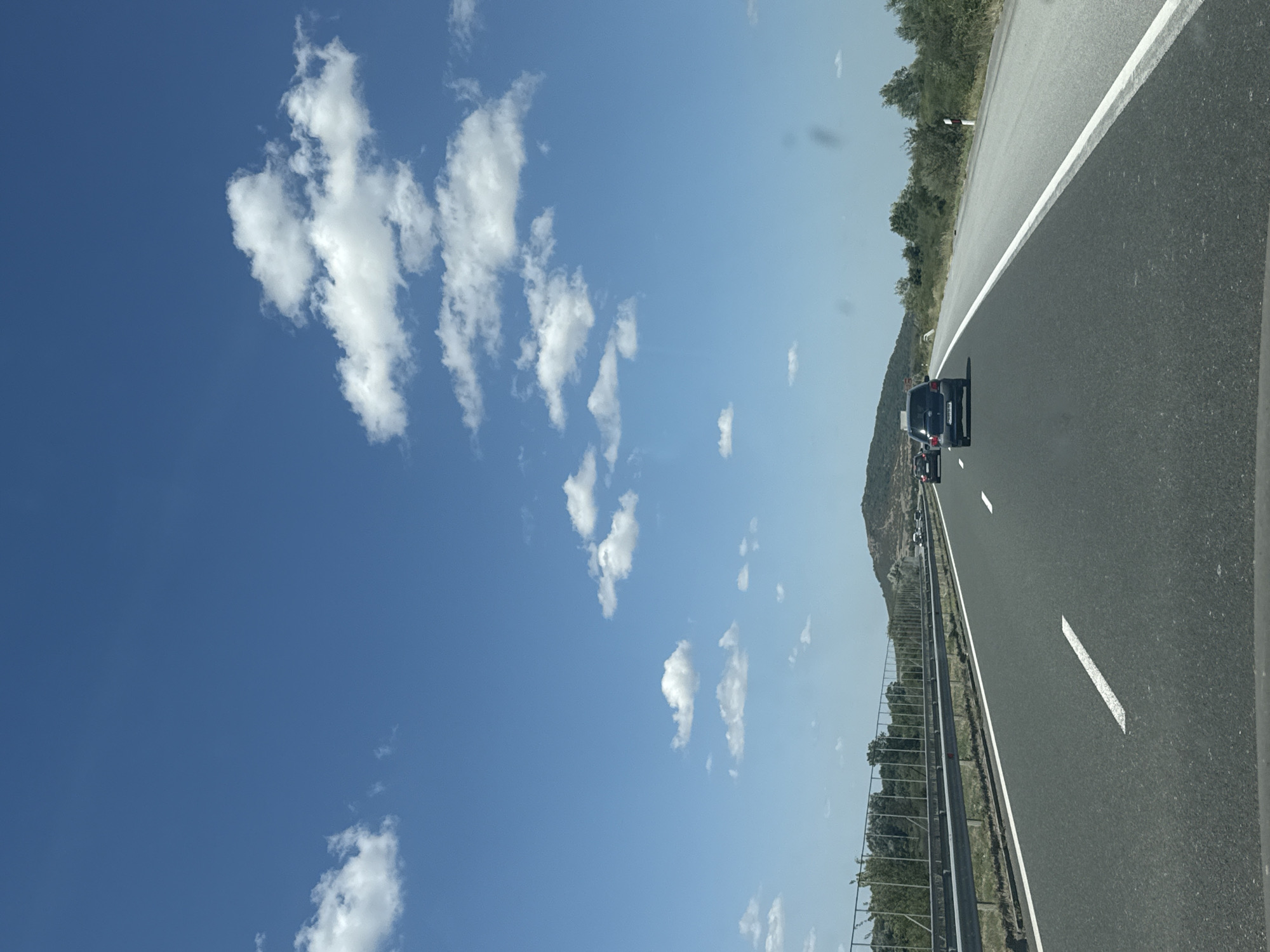 The image depicts a view from a vehicle traveling on a highway with a clear blue sky dotted with white clouds. The road has multiple lanes separated by white lines, and a metal guardrail runs along the left side, separating the highway from a grassy embankment and vegetation. In the distance, a hill or mountain is visible under the expansive sky. Several cars are on the highway, with a black SUV in the foreground and a few other vehicles further ahead. The environment appears to be rural or semi-rural, with trees and scrubland bordering the road. The image is taken during daylight hours, suggesting fair weather. The location is identified as Zavoy, Bulgaria.