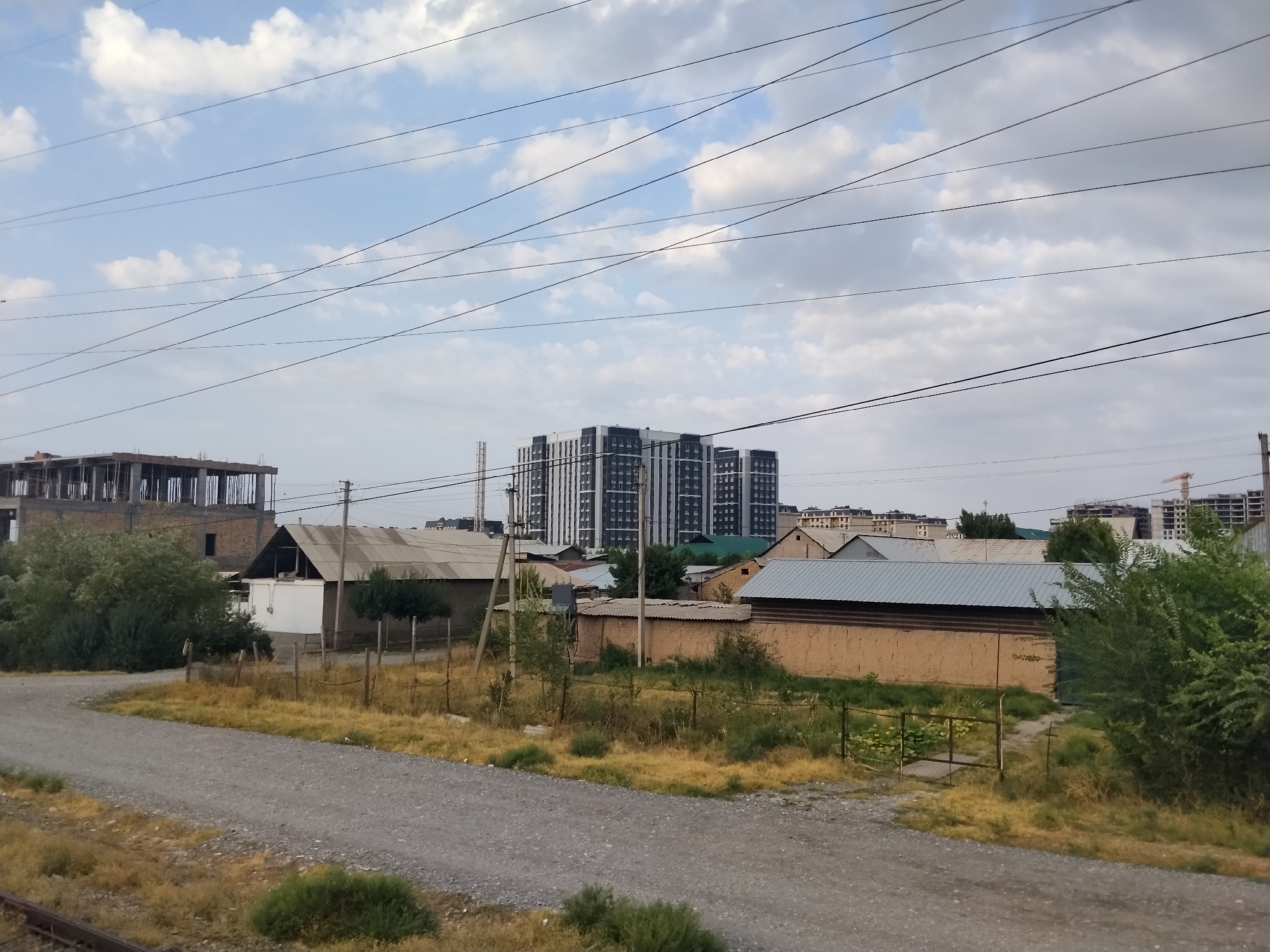 The image captures a view of a semi-urbanized area with a mix of older, low-rise structures and modern high-rise buildings in the background. In the foreground, a gravel road leads into the scene, bordered by dry, yellowed grass and sparse vegetation. A wire fence runs along the side of the road, enclosing several simple, single-story buildings with corrugated metal roofs and mud-brick walls. One building has a white facade with a dark, sloped roof. A partially constructed building with exposed rebar and concrete pillars is visible on the left. In the mid-ground, a cluster of apartment buildings with a distinct black and white facade stands out against a cloudy sky. Beyond these, other residential buildings and construction sites are discernible. Power lines crisscross the sky, and a utility pole is present near the fenced area. The overall impression is one of ongoing development and a juxtaposition of old and new infrastructure. The location is identified as Manas, Kyrgyzstan.