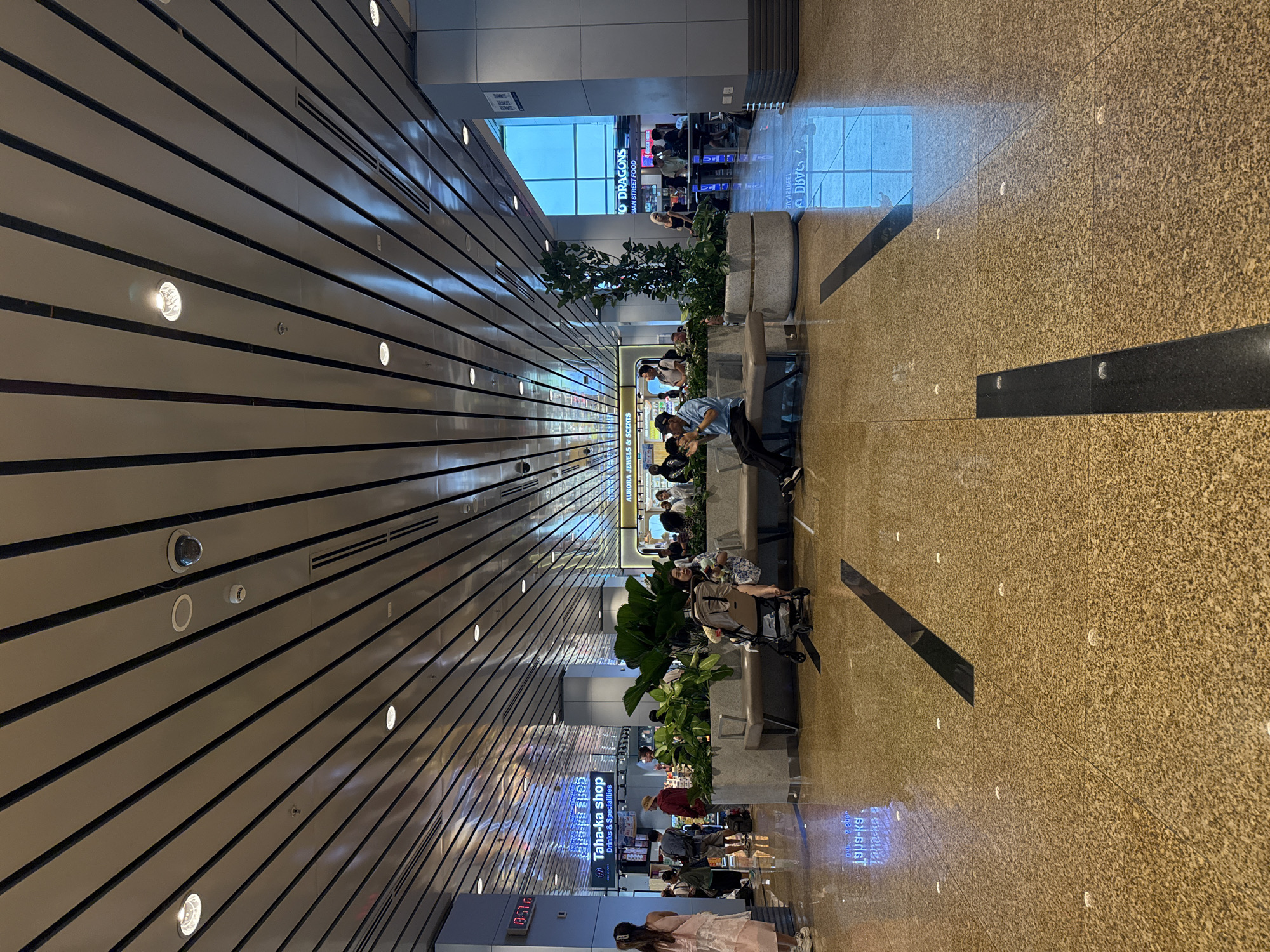 This image shows a public indoor space, likely an airport terminal, with a modern architectural design. The ceiling features a slatted design with integrated lighting and air vents, and several security cameras are visible. The floor is a polished speckled terrazzo material.
In the foreground, a person is seated on a bench, and a stroller is next to them. Several other people are visible in the mid-ground, some sitting on benches and others standing. The individuals are dressed in casual clothing.
There are several commercial establishments visible in the background, including a shop labeled "Taha-ka shop Drinks & Specialties" and another with signage that appears to be "AURORA JEWELS & SCENTS." Further down, there is a food establishment with the sign "TO DRAGONS ASIAN STREET FOOD." Large potted plants are strategically placed throughout the area, adding greenery to the environment. Reflections of the overhead lights and ceiling are visible on the polished floor.
The overall impression is a clean, well-lit, and functional public space designed for transit and commercial activity. The location is identified as Bắc Cam Ranh, Vietnam, which suggests it is within Cam Ranh City, Khánh Hòa Province, Vietnam, likely Cam Ranh International Airport.