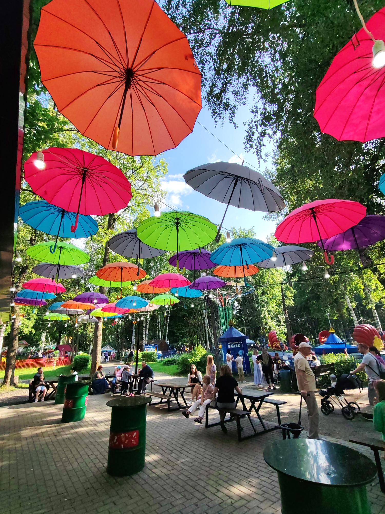 The image depicts a park scene in Izhevsk, Russia, characterized by a vibrant canopy of colorful umbrellas suspended overhead. The umbrellas, in shades of orange, pink, blue, green, and purple, are strung across the pathways, creating a festive and shaded atmosphere. In the foreground, people are seen gathered around picnic tables and green cylindrical trash receptacles. Some individuals are seated at the tables, while others are standing or pushing strollers. The background reveals a wooded area with trees, and hints of amusement park rides are visible. The overall impression is that of an outdoor recreational space, likely a public park or a fairground, adorned for an event or as a permanent decorative feature.