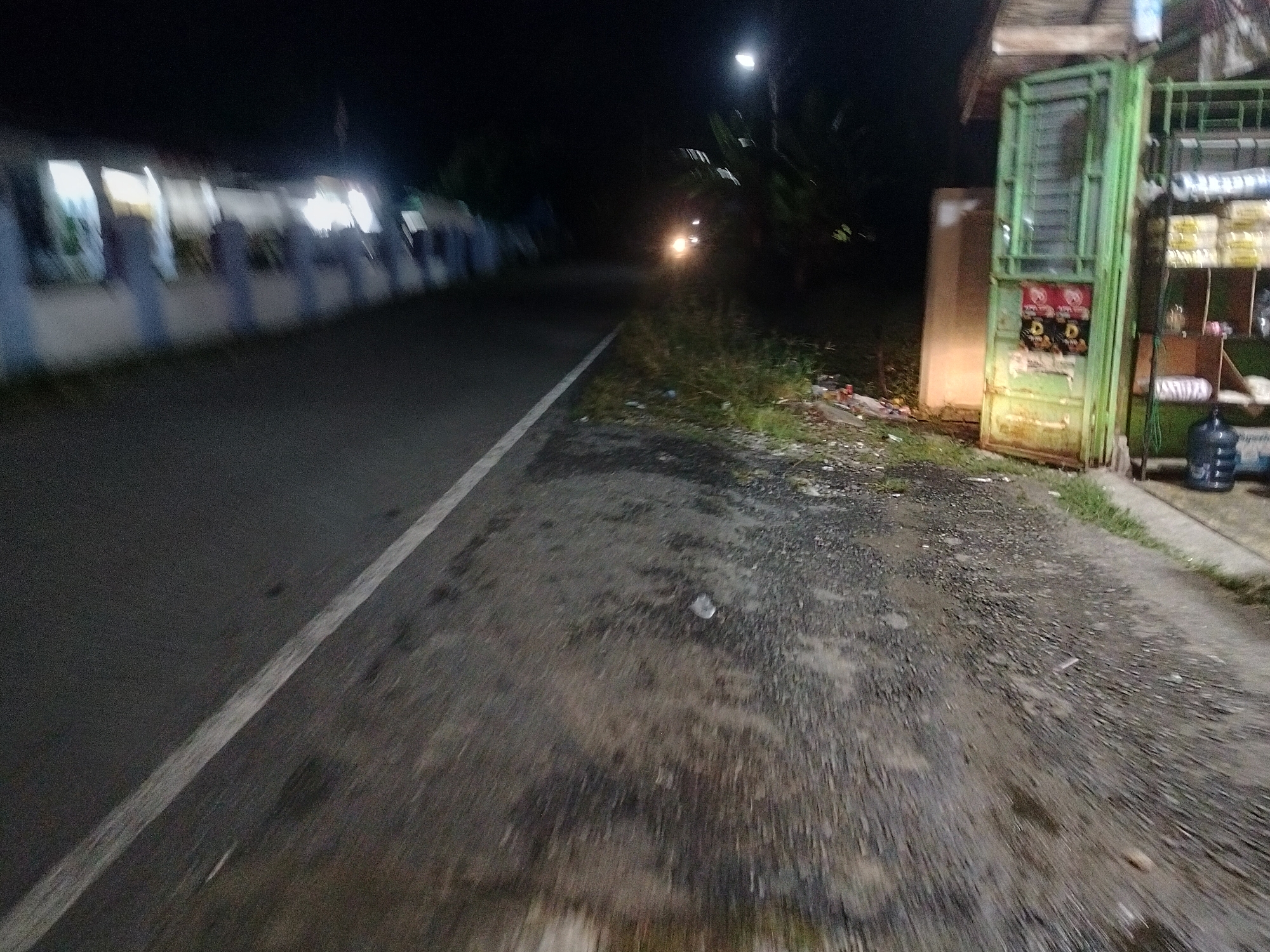The image depicts a dimly lit street scene at night in Kumbangindah, Indonesia. On the left side of the road, a long, low building with a white facade is visible, bordered by a white and blue railing. The road itself is dark asphalt with a white line marking the edge. To the right of the road, there is a small, open-fronted shop with a green metal frame and shutters. Shelves are visible inside the shop, displaying various items, and a large blue water jug sits outside. The ground beside the road is covered in gravel and grass, with some scattered debris. A single headlight from an approaching vehicle is visible in the distance, illuminating the road ahead. The overall atmosphere is nocturnal and somewhat deserted, with artificial lighting from the building and the shop casting faint glows.