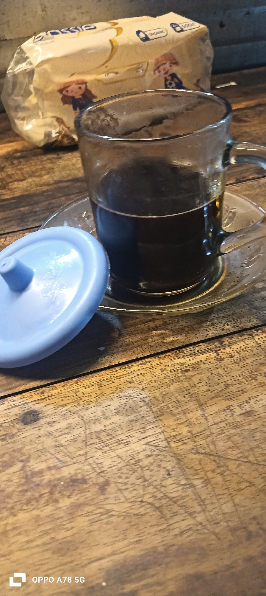 A close-up shot shows a glass mug filled with dark liquid, likely coffee, placed on a wooden table. Next to the mug is a pale blue lid with a small opening. In the background, partially visible, is a crinkled package with illustrations of children and the word "ORGANIC". The wooden surface displays a textured grain and some scratches. The bottom left corner of the image shows a watermark reading "OPPO A78 5G". The scene appears to be indoors, possibly in a cafe or home setting.