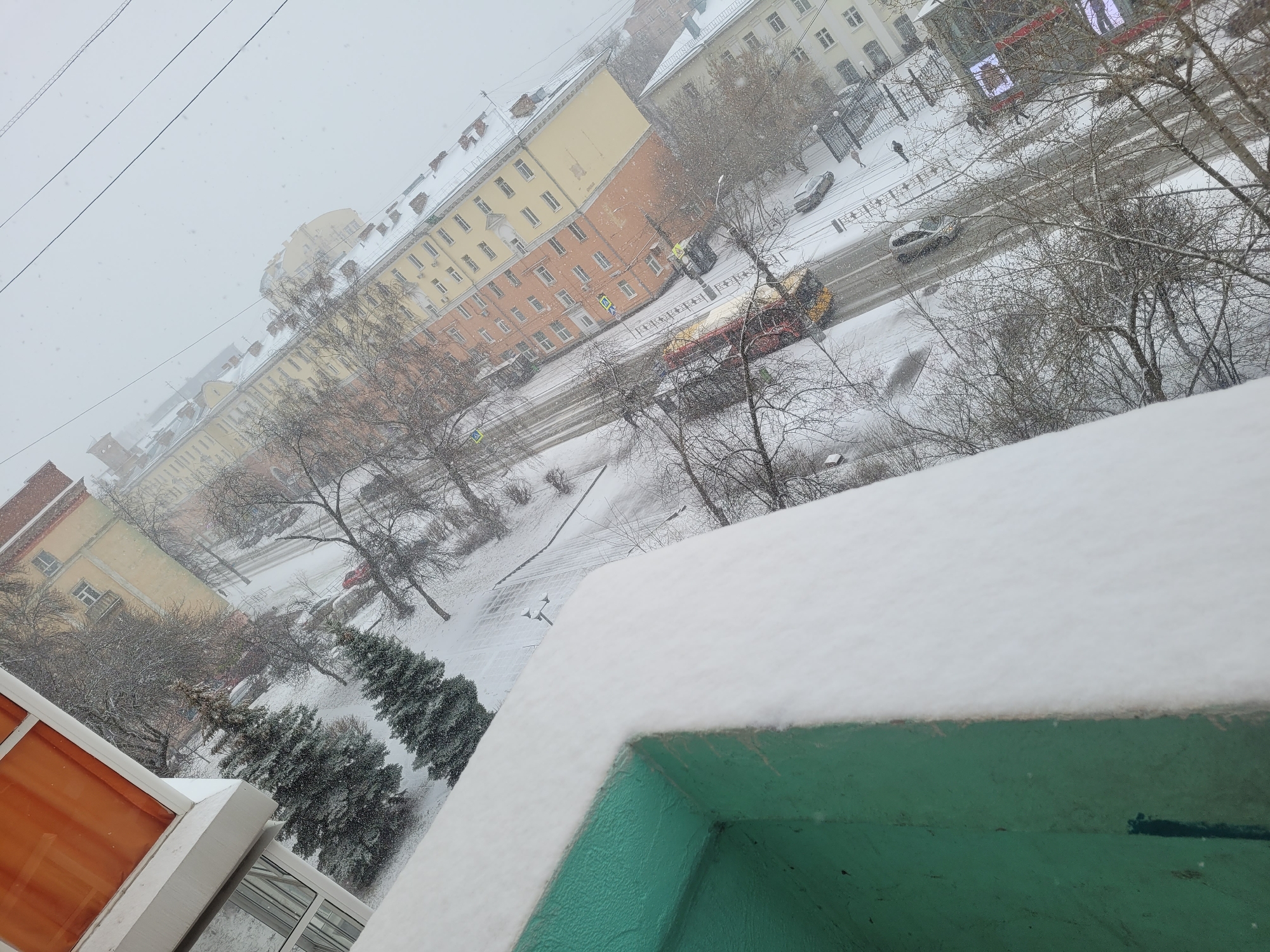 The image depicts a snow-covered urban scene from an elevated perspective, likely from a balcony or high window. In the background, buildings of varying heights and architectural styles line the streets, also covered in snow. Bare trees are scattered throughout the scene. In the street, there are vehicles visible: cars and what appears to be a truck or bus. The foreground is dominated by the edge of the balcony, coated in snow, with its blue-green surface visible. In the lower left corner, a portion of an orange window frame is visible.
The location is confirmed to be the city of Izhevsk, Russia.