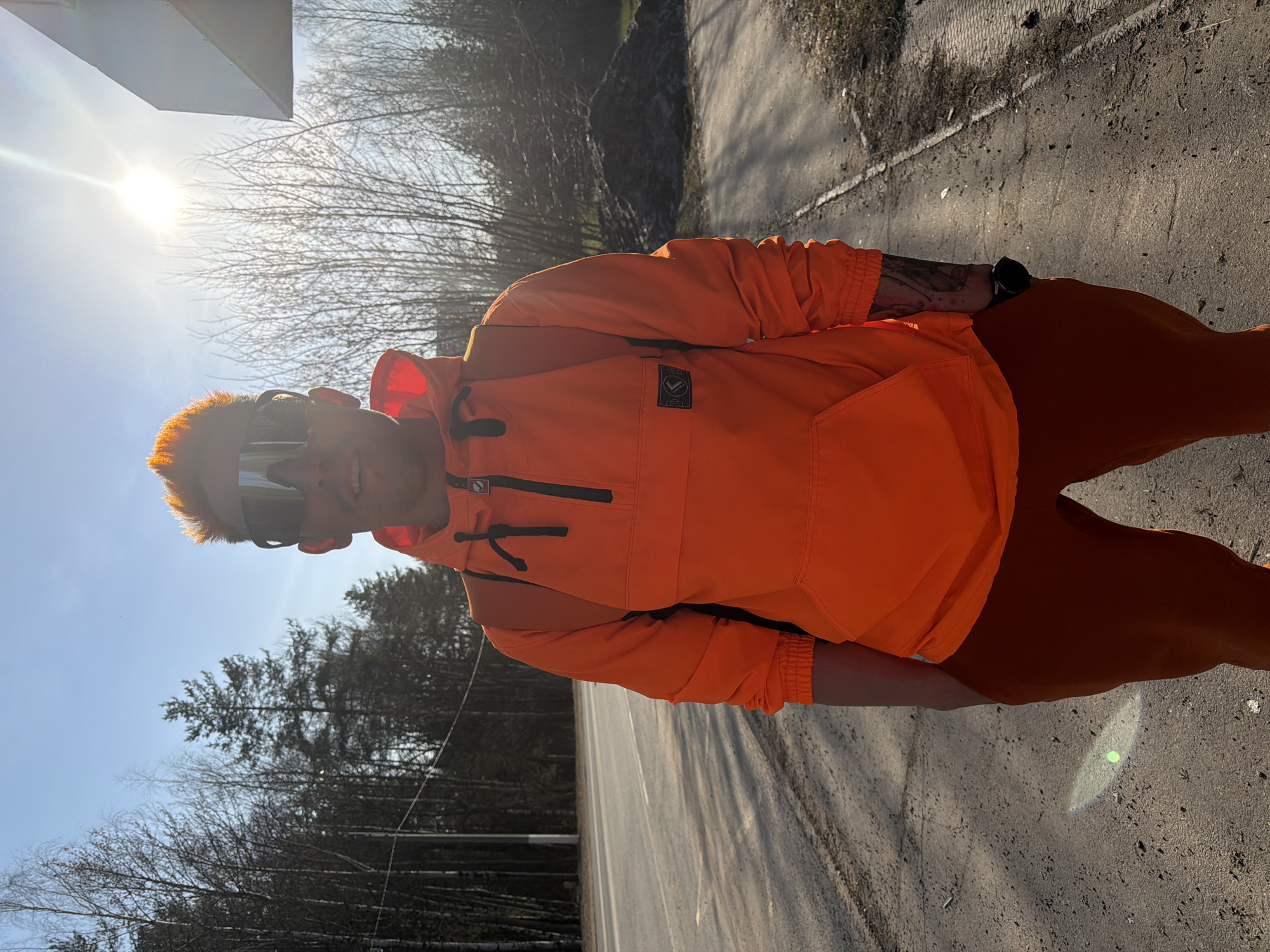 A person wearing orange clothing and sunglasses stands on a gray asphalt road next to a wooded area under a bright, sunny sky. The trees behind them are bare and silhouetted against the sunlight. The location is identified as the city of Chaikovsky, Russia.