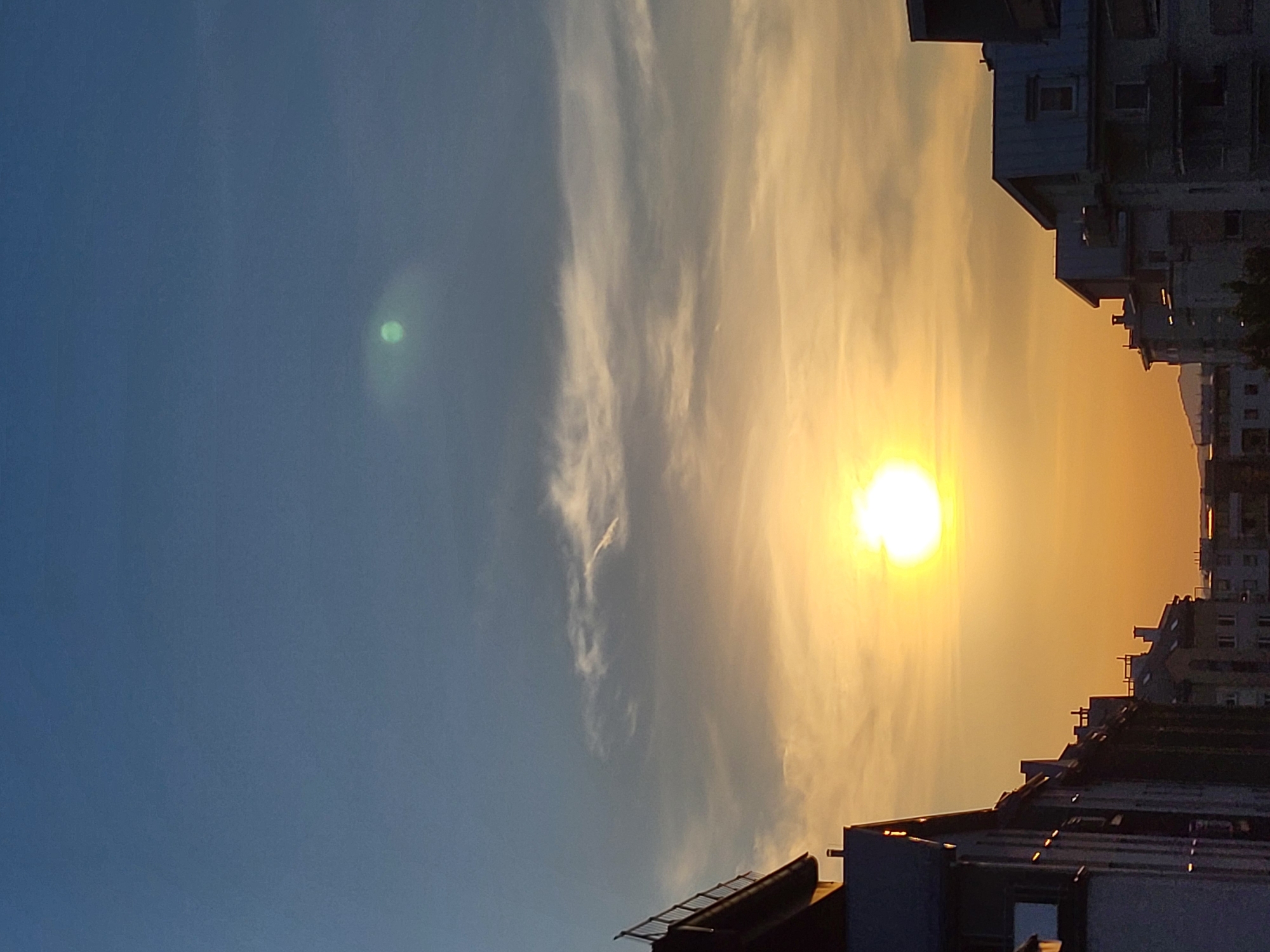 The image captures a view of a city skyline at sunset. The sky is a gradient of deep blue at the top transitioning to warm oranges and yellows near the horizon. A bright, large sun is visible, casting a strong glow and creating a haze around it. In the upper portion of the sky, there is a faint, greenish circular anomaly, possibly a lens flare or atmospheric phenomenon. Wispy clouds are scattered across the sky, some illuminated by the setting sun.
Silhouetted buildings line the bottom of the frame, their architectural details muted by the low light. The buildings appear to be of varying heights and styles, with some featuring pitched roofs and chimneys. The perspective suggests the photo was taken from a higher vantage point, looking down or across the rooftops. The scene is devoid of any people or visible vehicles. The overall atmosphere is serene and captures the beauty of a fading day in an urban environment.
