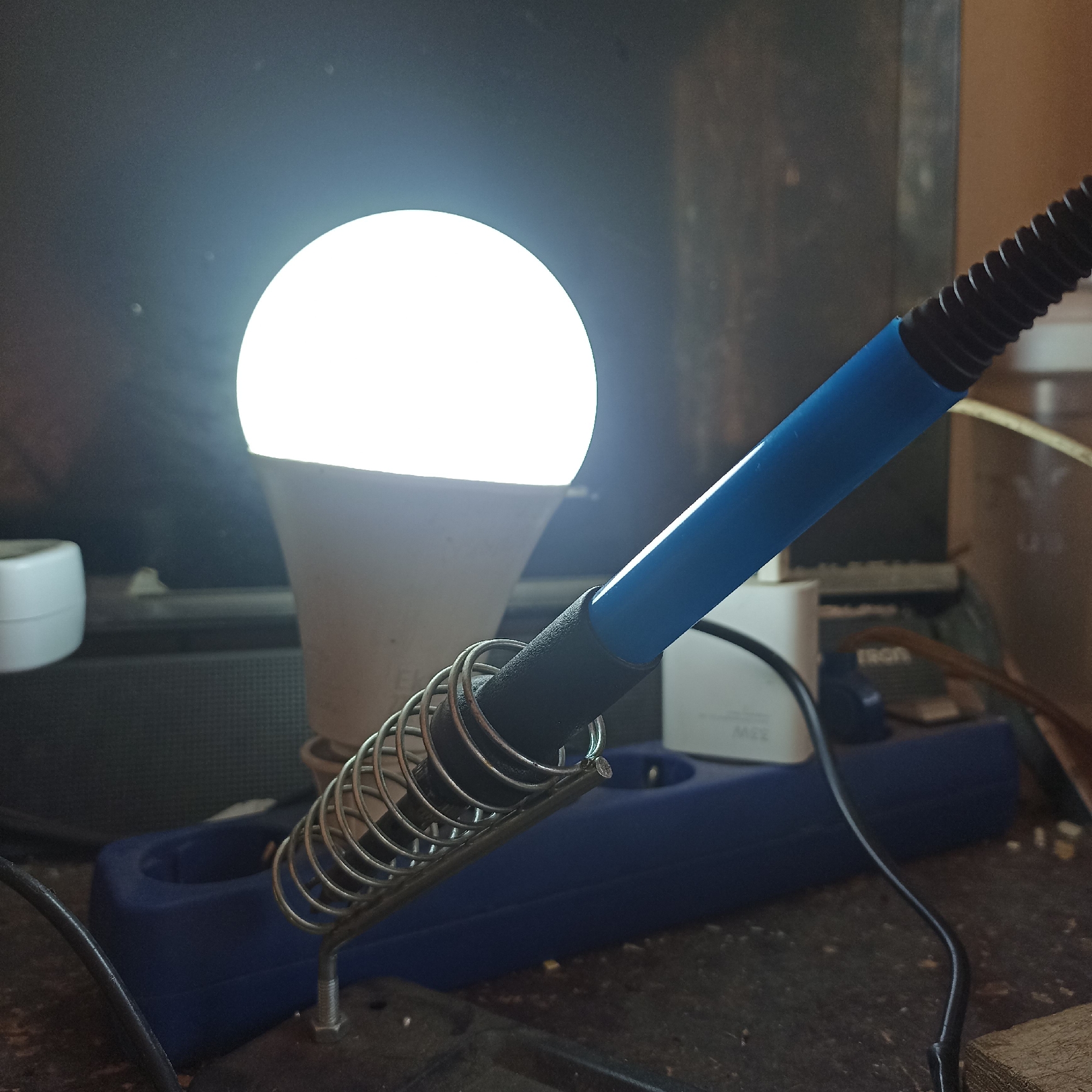 The image shows a close-up of a soldering iron resting in its stand. The soldering iron has a blue handle and a black coiled cord. The stand is made of metal and is placed on a dark, textured surface, possibly a workbench. In the background, a bright LED light bulb is illuminated, casting a strong light. Behind the light bulb, a dark object, possibly a computer monitor, is visible. To the left of the light bulb, a white cylindrical object, likely a plug or adapter, is partially visible. A blue power strip with electrical outlets is situated behind the soldering iron stand. The scene appears to be an indoor workshop or electronic repair area. There is no indication of the city of Magelang, Indonesia.