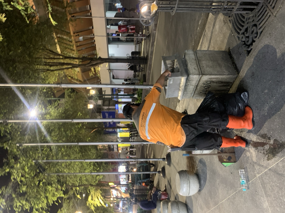 At night in Yogyakarta, Indonesia, a male sanitation worker is observed on a brightly lit sidewalk. He wears an orange long-sleeve shirt with "SODOBORO" and additional text, black cargo pants, bright orange rubber boots, a dark baseball cap, and a black face mask. His back is mostly to the viewer as he empties the contents of a grey square public trash receptacle, mounted on a stone base inscribed with "jogja," into a black trash bag positioned on the ground next to his left boot. A blue plastic water bottle lies horizontally on the sidewalk near his right foot, which is also close to a long-handled brush or broom.
The sidewalk features multiple light-colored, spherical concrete bollards and a decorative cast-iron grate for drainage. Directly behind the worker, a row of tall, thin silver poles extends into the background. Lush green trees, illuminated by streetlights, line the thoroughfare. In the background, other individuals are visible on the sidewalk and street, which appears busy with parked motorcycles, including a three-wheeled vehicle on the left, and brightly lit storefronts with various signs, one partially legible as "TONGCHAT." Buildings with distinctive brown-patterned facades are visible further down the street.