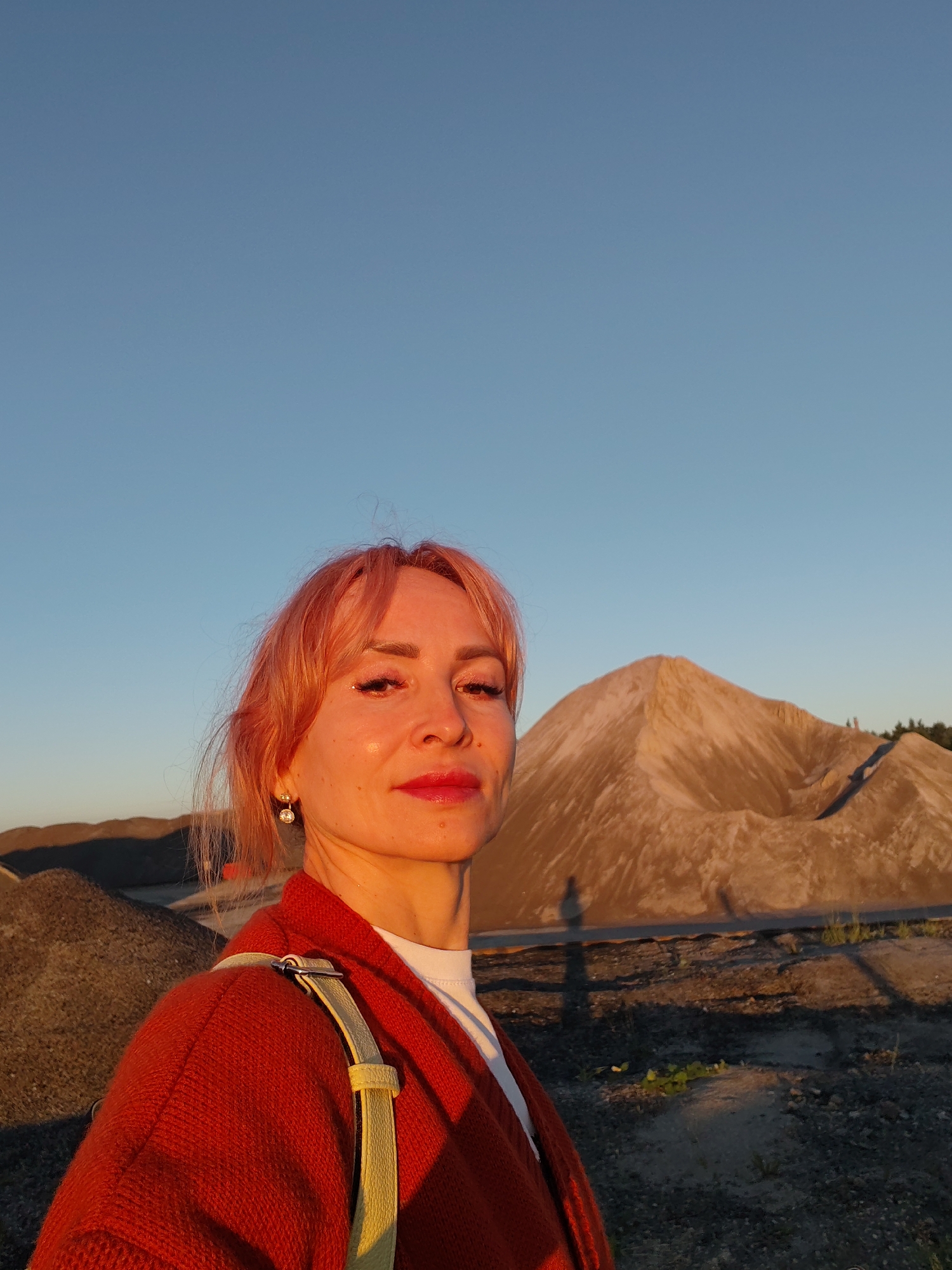 A woman with pink hair takes a selfie outdoors during golden hour, with a bright blue sky above. She is wearing a red knitted cardigan over a white top and a light yellow strap from a bag is visible over her shoulder. She has on bright red lipstick and subtle glittery eye makeup. She is also wearing a pair of small, sparkly earrings.
In the background, there are large piles of gravel or sand, one of which forms a prominent mound that is lit by the setting sun, casting long shadows. The ground is a mix of sand and gravel, with some sparse vegetation visible. The scene appears to be a quarry or industrial site. The time of day is clearly sunset due to the warm, golden light bathing the landscape and the long shadows. The weather appears to be clear and fair. The location is identified as Bol'shie Mkhi, Russia. No text is visible in the image. The woman's expression is neutral and she is looking directly at the camera.