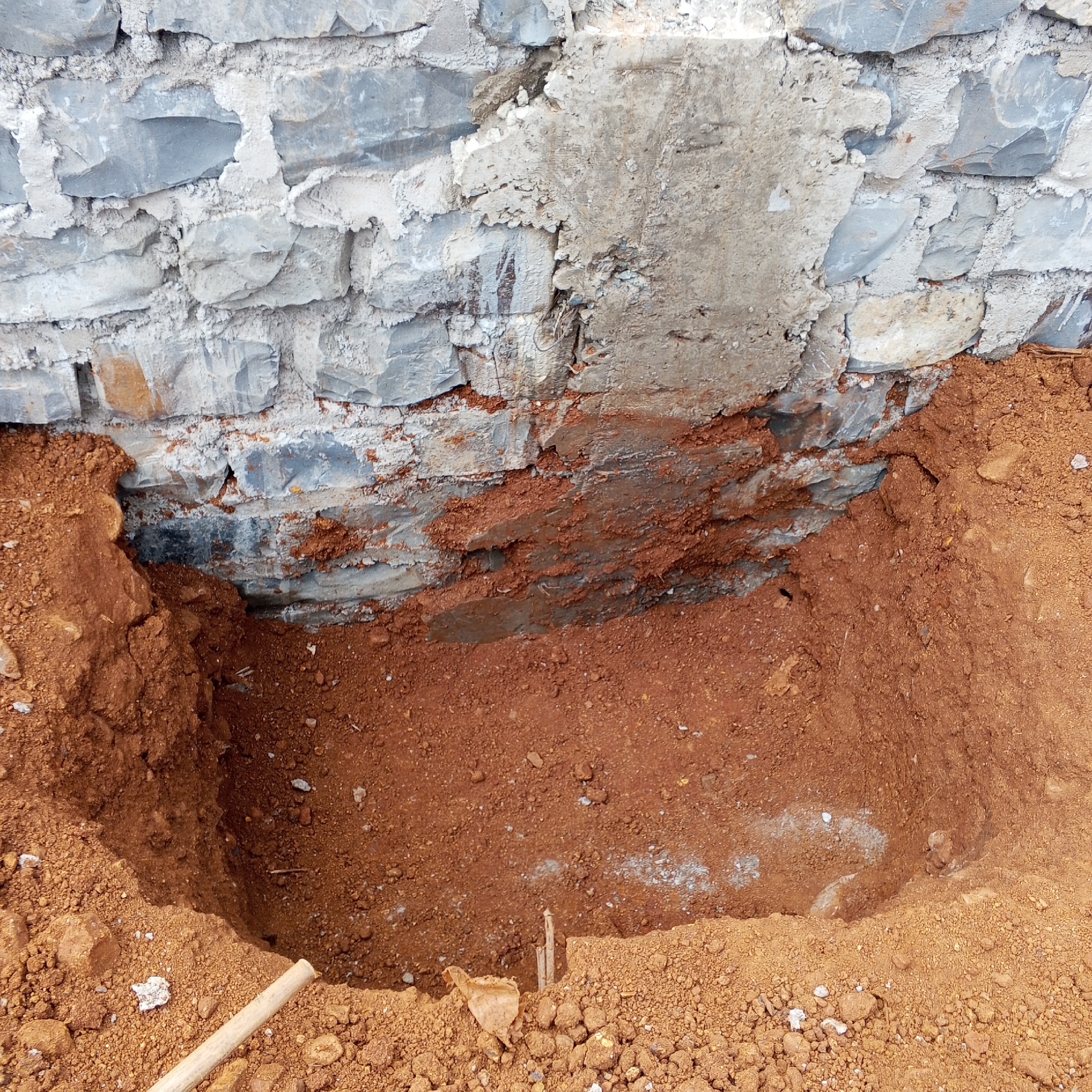 The image depicts a hole dug in the ground next to a stone foundation wall. The soil is reddish-brown. The stone wall is made of gray and white stones and mortar. There is a patch of exposed concrete on the wall. A wooden handle is visible in the lower left corner of the hole. Two sticks are in the hole. The image may have been taken in the Borabu Chitago ward of Kenya.