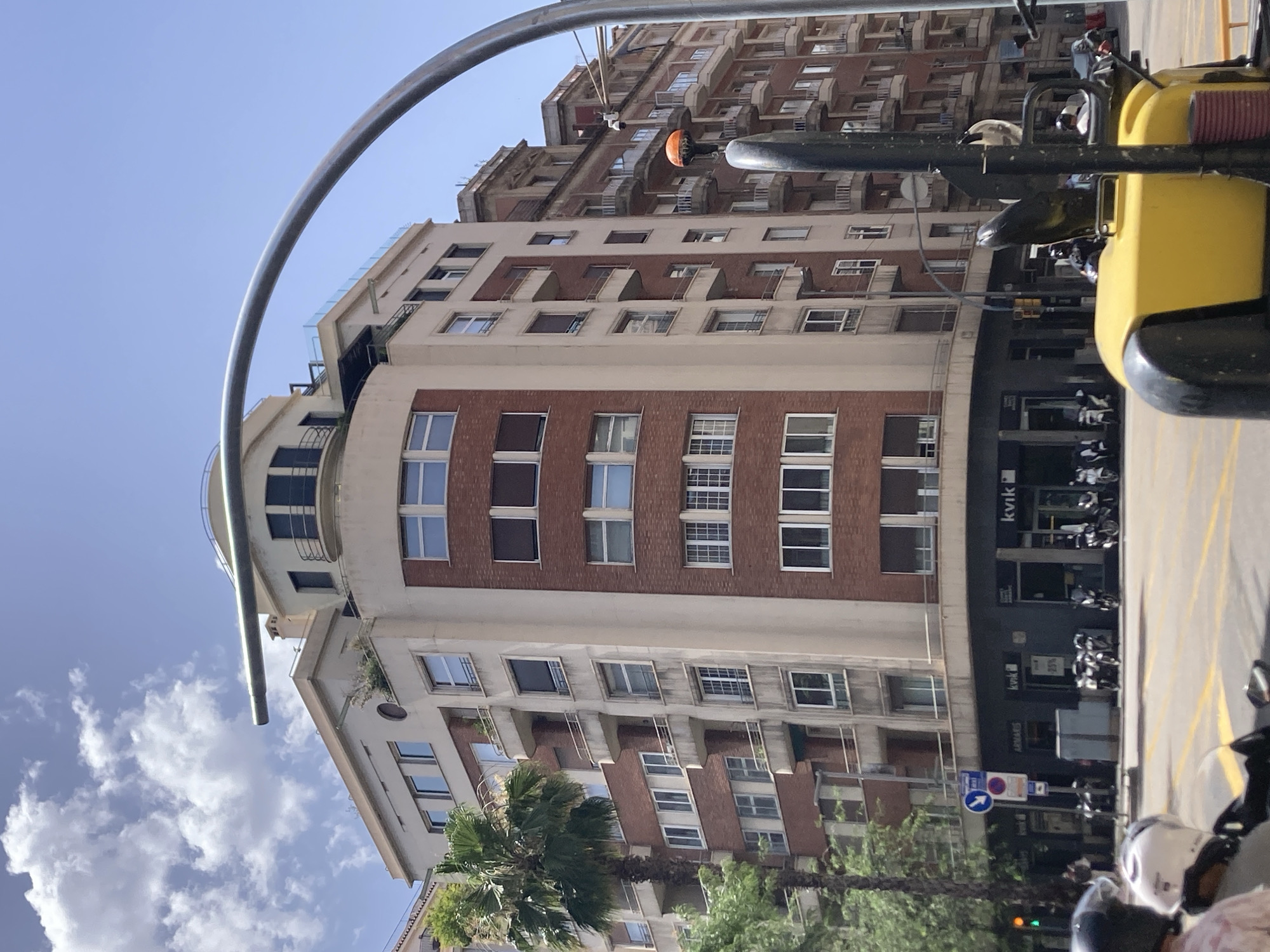 The image shows a street scene in Barcelona, Spain. A modern building with red brick and cream-colored sections dominates the center of the frame, featuring multiple floors with balconies and numerous windows. A tall palm tree stands to the left of the building. In the foreground, part of a yellow construction vehicle is visible, along with the backs of two people on motorcycles wearing helmets. Several parked motorcycles are lined up along the sidewalk in front of shops on the ground floor of the building. Traffic lights and street signs are also present. The sky is blue with scattered clouds.