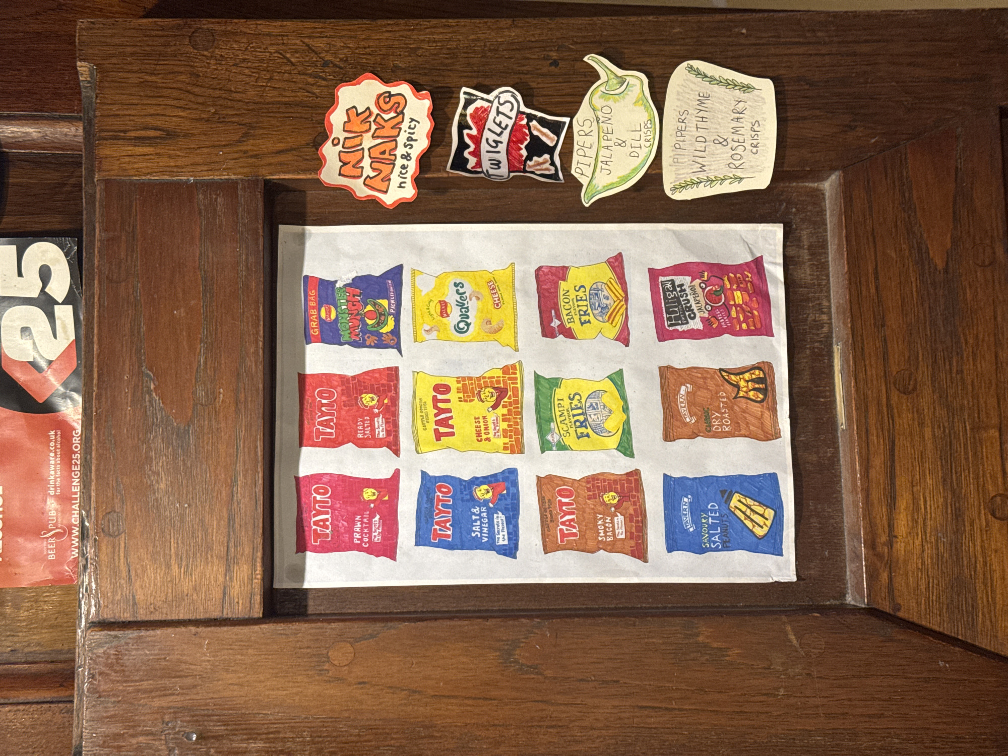 The image shows a wooden-framed noticeboard, likely inside a pub or bar. Attached to the board are various stickers, including "Nik Naks nice & spicy", "Twiglets", "Pipers Jalapeno & Dill Crisps", and "Pipipers Wild Thyme & Rosemary Crisps". Below the stickers is a drawing on white paper, depicting various brands and flavors of crisps (potato chips), including "Monster Munch", "Quavers", "Bacon Fries", "Huligan Crush Jalapeno Pretzel", and several varieties of "Tayto" crisps. A magazine or promotional flyer for "Beer Pub" and the "Challenge25" campaign is partially visible on the left edge of the frame. The entire scene is mounted on a wooden surface.