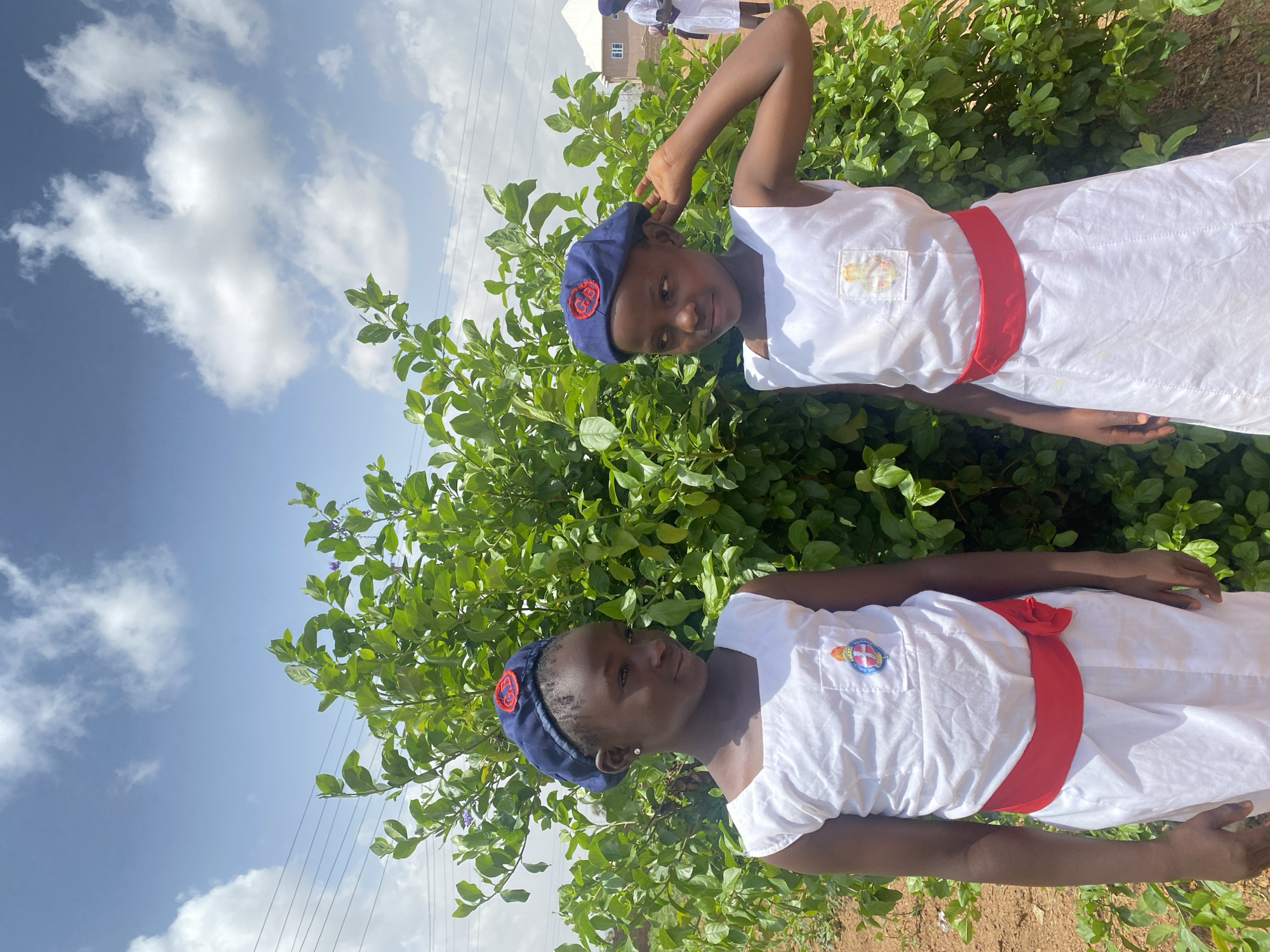 Two girls stand side-by-side in front of a leafy green shrub, against a backdrop of a light blue sky with scattered clouds. Both girls wear matching white dresses with a red sash around the waist and blue caps. A building can be partially seen in the background behind the shrub. The location is Turu, Nigeria.