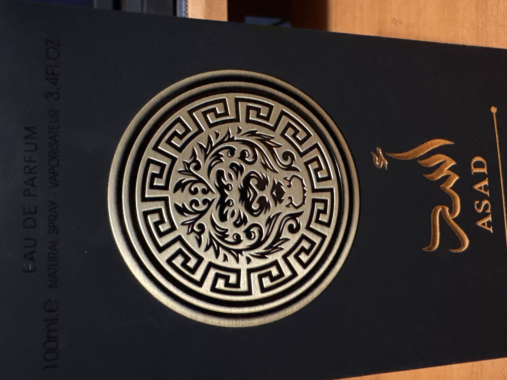 A dark, rectangular product box, likely for perfume, is prominently displayed in the image. The box has a matte, black or very dark blue finish.
A large, circular, gold-colored emblem is centered on the box. This emblem features an intricate, ornate design in its core, resembling stylized foliage or a mythical creature. Surrounding this central motif is a distinct Greek key (meander) pattern, forming the outer border of the circular emblem. The gold elements of the emblem appear to be embossed, providing a tactile texture and metallic sheen.
Text is vertically printed on both sides of the box. On the left, in a light gray or silver font, the following is visible: "100ml.e EAU DE PARFUM NATURAL SPRAY - VAPORISATEUR 3.4FL.OZ". On the right side, gold text is visible, reading "ASAD", positioned below a stylized golden design that appears to be Arabic script, likely the brand's name. Both the "ASAD" text and the Arabic script are also embossed or foiled in gold.
The image is captured at a slight clockwise angle. The background is softly blurred, revealing a warm, wooden surface, possibly a table, and some indistinct darker objects. The lighting suggests an indoor setting. There is no activity taking place; the image is a static product shot. The combination of the dark box and gold accents suggests a premium or luxury item.