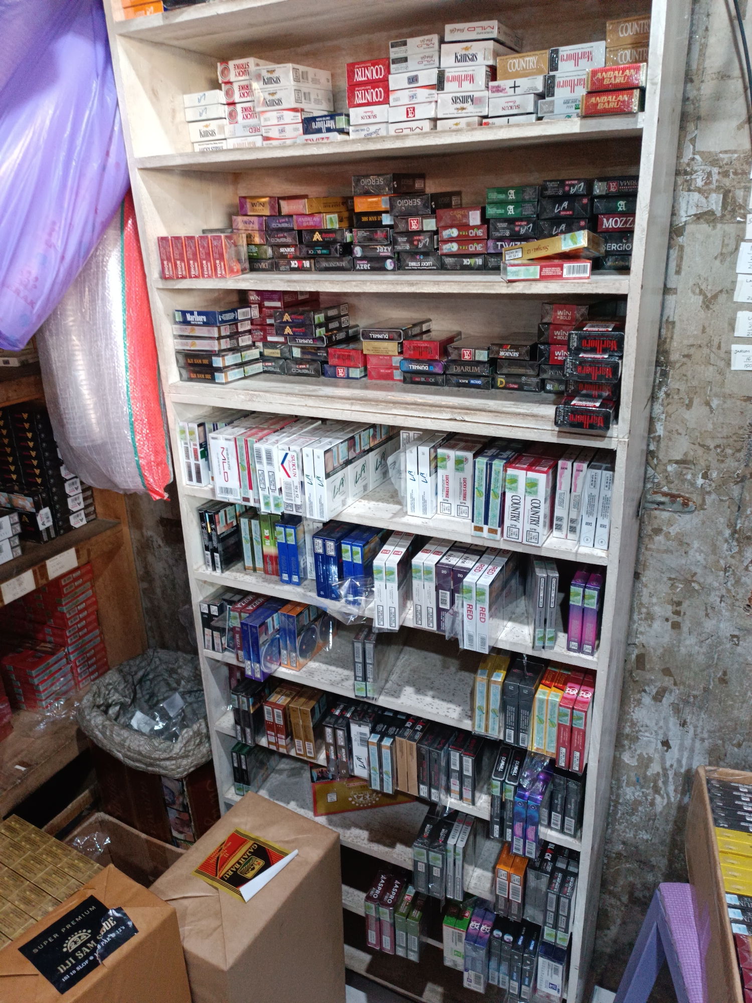 This image depicts the interior of a small convenience store or "warung" in Yogyakarta, Indonesia. The store is packed with shelves filled with various brands of cigarettes. The shelves are made of light-colored wood and are stacked from floor to ceiling. On the left side of the image, large plastic-wrapped items, possibly imported goods or large rolls of fabric, are leaning against the wall.
The merchandise is predominantly cigarette packs, neatly arranged in rows. Prominent brands visible include Marlboro, Country, Kansas, Lucky Strike, LA, Dunhill, and others, often displayed in their original cardboard packaging. Some shelves also contain other miscellaneous items, such as bags of what appears to be snacks or dry goods, and a large plastic bag containing small items.
In the foreground, on the right, a wooden structure, possibly a display stand, holds more cigarette packs. A small, purple, stool-like object is positioned near it. The walls of the store appear to be made of rough, textured concrete or brick. The lighting suggests an indoor environment, possibly with fluorescent lights overhead, although they are not directly visible. The overall impression is of a densely stocked retail space common in Indonesian local markets.