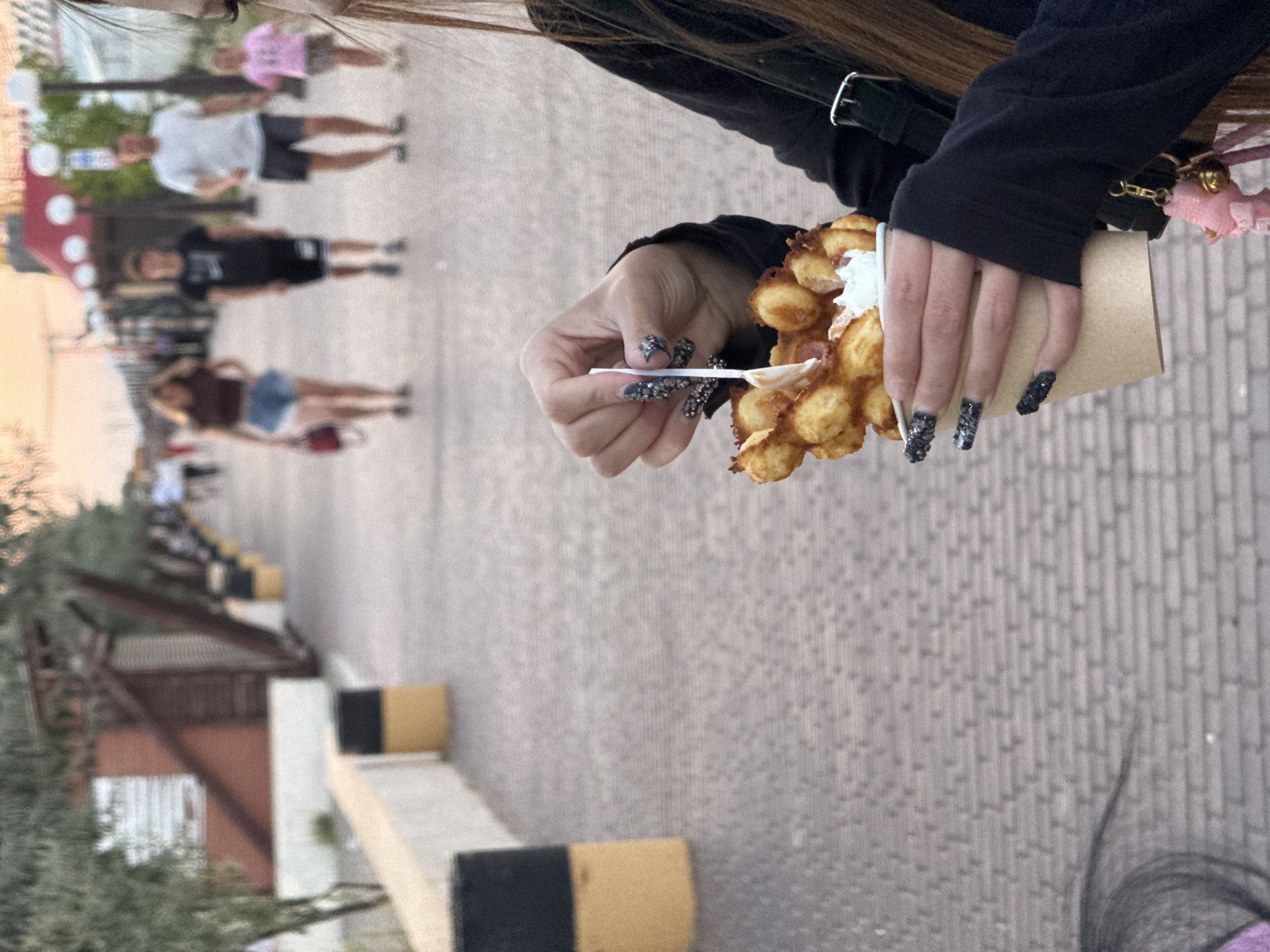 A close-up shot focuses on a person's hand holding a bubble waffle in a paper cup. The waffle is golden brown and appears to have some filling or topping. The person's nails are long and decorated with dark glitter. The background is blurred but suggests an outdoor environment, possibly a boardwalk or promenade with other people walking in the distance. The ground is paved with bricks. There are also some trees and structures visible in the background, along with trash cans. The lighting suggests it is late afternoon or early evening. The scene does not appear to be located in Novofedorovka, Ukraine, as there are no specific visual cues to indicate this location.