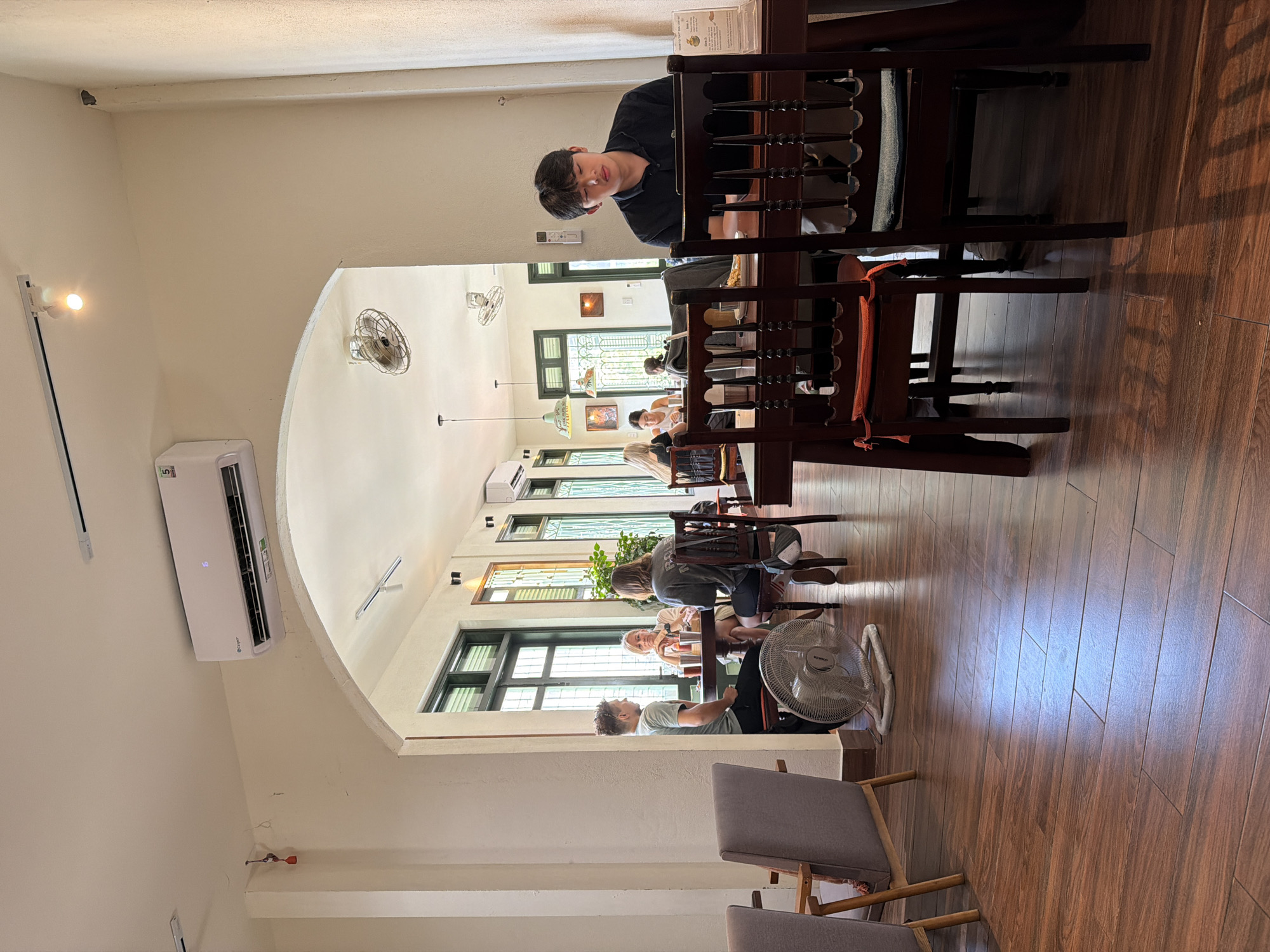 The image depicts the interior of a cafe, possibly in Da Nang, Vietnam, characterized by its warm, earthy tones and natural lighting. The flooring is made of dark wooden planks, reflecting the ambient light. In the foreground, a dark wooden table with ornate chairs is visible. On the right, a young Asian man in a dark shirt is seated at the table, looking down at a laptop, with his eyes closed or lowered. A small white sign with text is on the table to his right.
In the mid-ground, an archway opens into another section of the cafe. On the left, a white standing fan is placed on the floor. Several people are seated at tables in this area. A man and two women are at a round table, engaged in conversation or eating. The man is wearing a grey t-shirt, and the women have blonde and brown hair. Further back, other patrons are visible at tables. The walls are painted in a light, neutral color, and architectural elements like columns and large windows with green frames are present. An air conditioning unit is mounted high on the wall above the archway.
The overall ambiance suggests a relaxed and casual setting, likely a popular spot for locals and tourists. The presence of plants and decorative elements in the background adds to the inviting atmosphere.