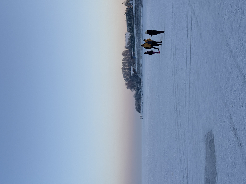 The image depicts an outdoor winter scene on a frozen body of water in Liutizh, Ukraine, under a clear, gradient sky transitioning from light blue overhead to a pale pink-orange near the horizon. The foreground and midground consist of a flat, extensive expanse of snow-covered ice, marked by linear tracks. In the lower-left foreground, an irregular, dark-toned patch, consistent with open water or thin ice, is visible. Four individuals are present in the midground, standing on the frozen surface in winter clothing. One individual wears a bright yellow jacket and dark pants, with one leg lifted, suggesting movement or preparation for ice skating. The background reveals a snow-covered shoreline featuring multiple bare trees, several buildings with visible roofs, and a low retaining wall. A short, dark-colored pier-like structure extends from the shoreline into the frozen water. Natural daylight illuminates the scene uniformly.