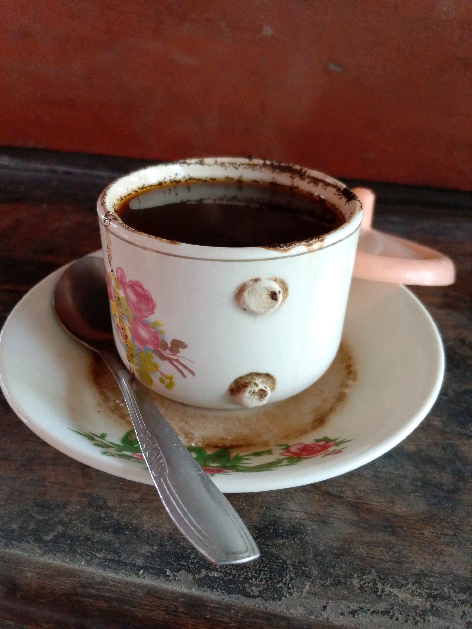 A white ceramic cup with a floral pattern containing dark coffee sits on a saucer on a wooden table. A silver spoon rests on the saucer. A pink object, possibly a lid, is visible behind the cup. The background is an out-of-focus reddish-brown surface. The scene appears to be indoors, likely in a home or cafe in Jiwan, Indonesia.