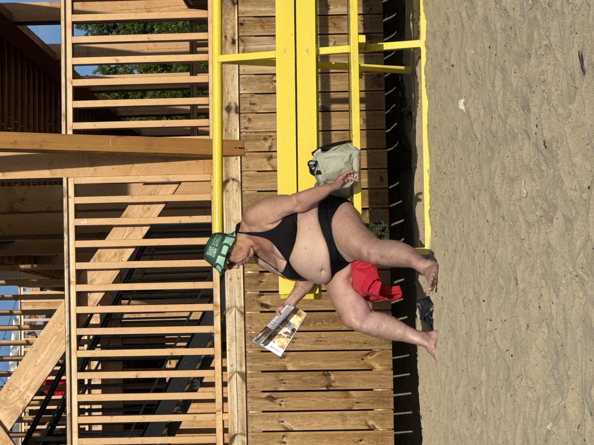 In a sandy outdoor area, likely a beach or park, a person wearing a black bikini and a green patterned bucket hat sits on a bright yellow bench. They are holding an open magazine in their hands and appear to be reading it. A red towel is draped over their lap, and a light-colored bag rests on the bench beside them. The background consists of a wooden structure with vertical slats and railings, suggesting a pier, boardwalk, or elevated deck. The ground is covered in sand, with shadows cast from the wooden structure. The lighting indicates a sunny day. No specific landmarks or indicators place this scene in Tallinn, Estonia.