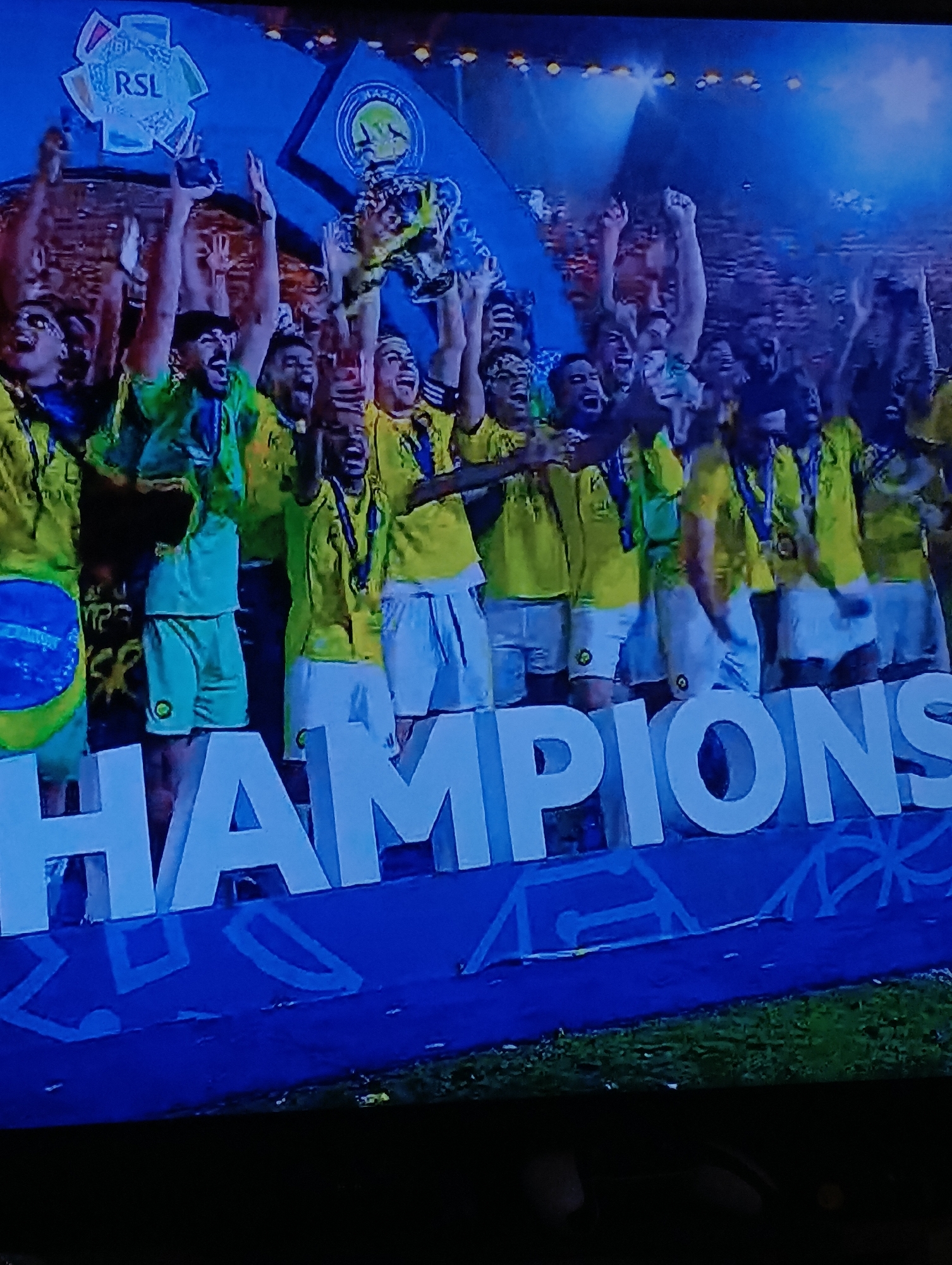 A group of people in yellow and green jerseys are celebrating behind large, white block letters spelling "CHAMPIONS." Many are holding their arms in the air. Some wear medals. There are "RSL" and "MAZZAA" logos in the background. The event appears to be taking place at a stadium in Plovdiv, Bulgaria.