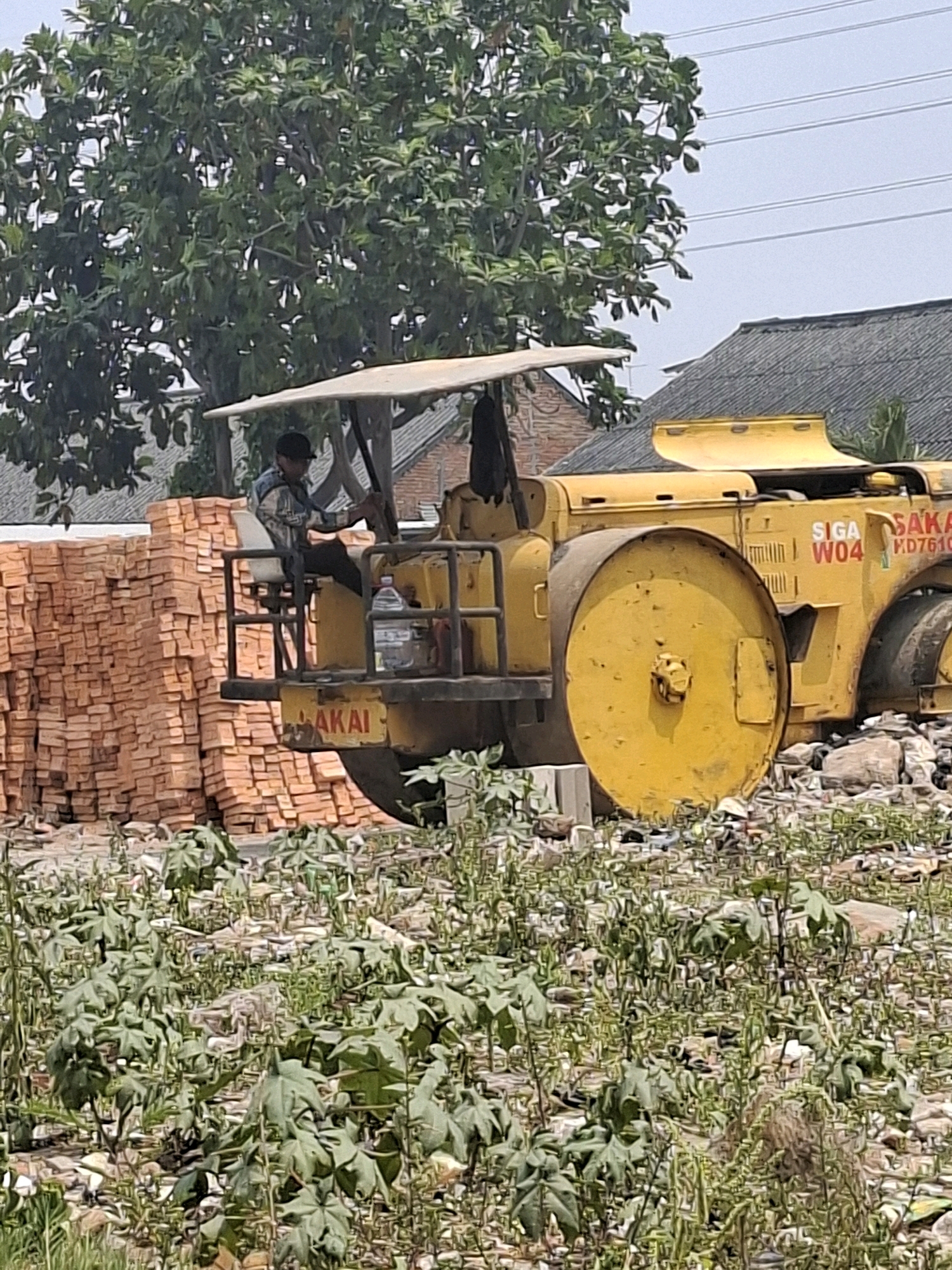 A yellow road roller is parked near a large stack of bricks. A person in a blue and white checkered shirt and a black cap is sitting in the operator's seat of the road roller. The road roller has the markings "SIGA W04 HD7610" on its side. A bottle of water is attached to the side of the operator's platform. The scene appears to be an outdoor construction or material storage area, possibly in a semi-urban or industrial part of North Jakarta, Indonesia, given the presence of building materials and a tree with large leaves. The ground is covered in debris and vegetation. In the background, there are buildings with tiled roofs and overhead power lines.
